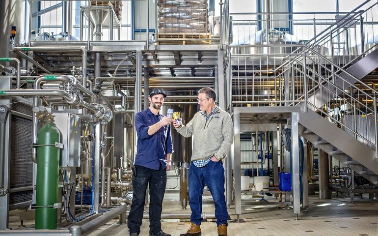 Rob Tod and Luke Truman on the Allagash Brewing factory floor, each holding a can of beer.