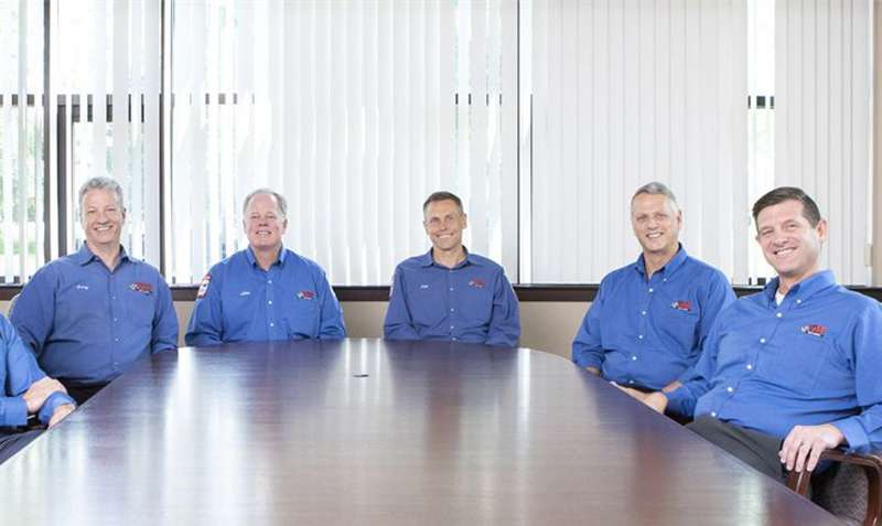 Five white men in bright blue shirts sit at a table facing the camera and smiling