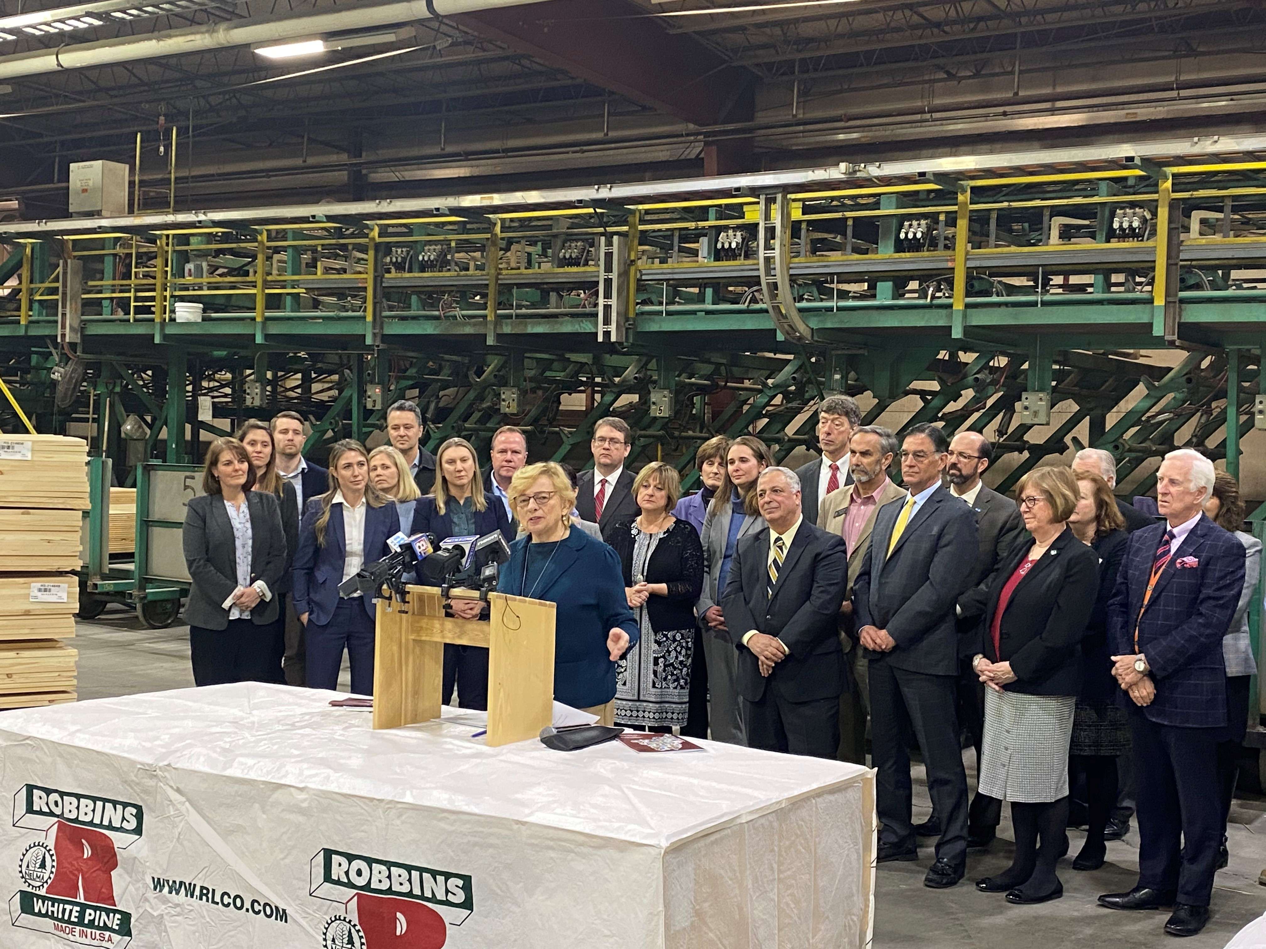 A woman speaks at a lectern as a about 20 people in business suits stand behind her. They are in a manufacturing plant.