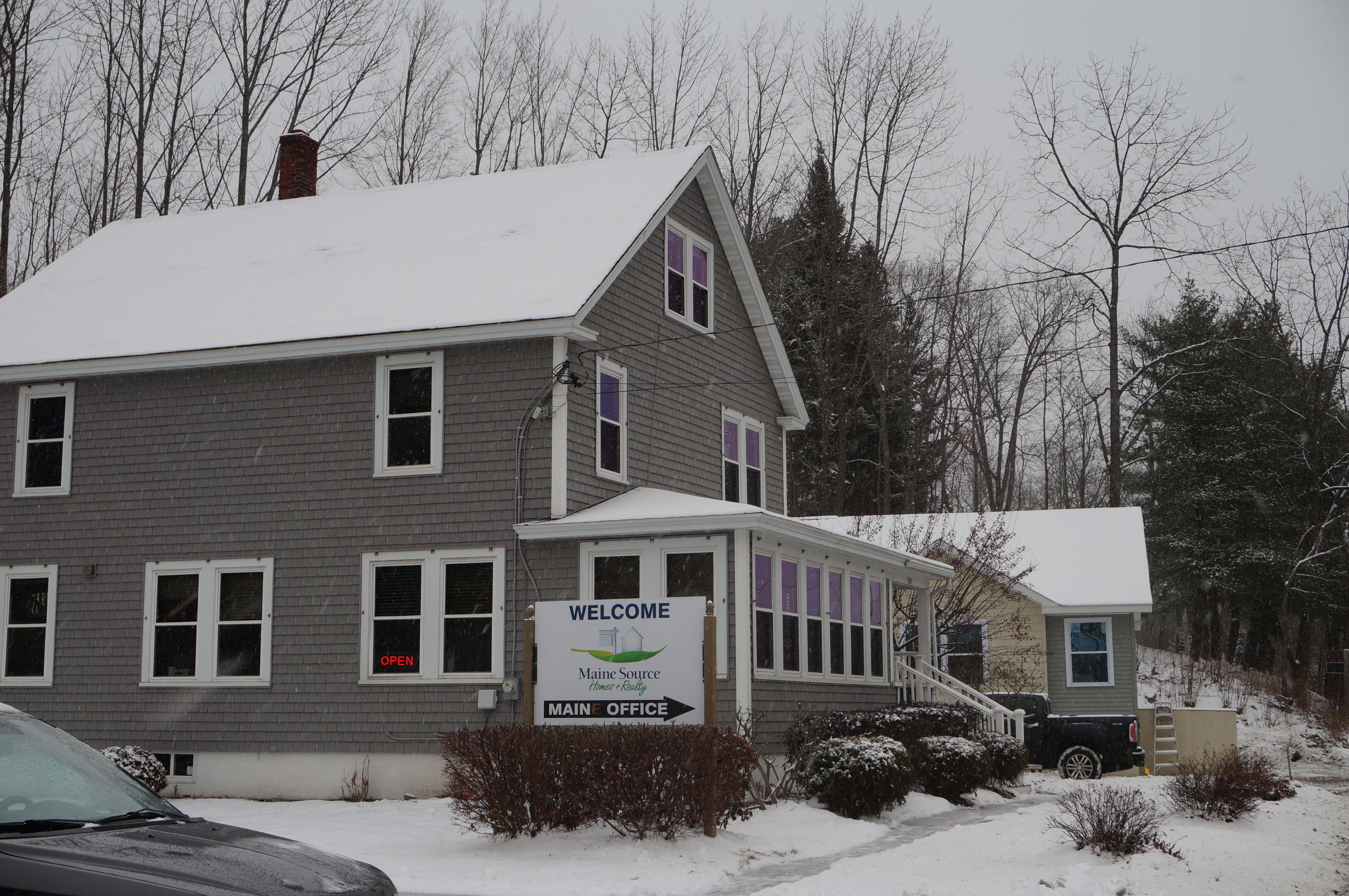 A two-story house with a sign that says Maine Source, and a neon "open" sign in the window on a snowy street