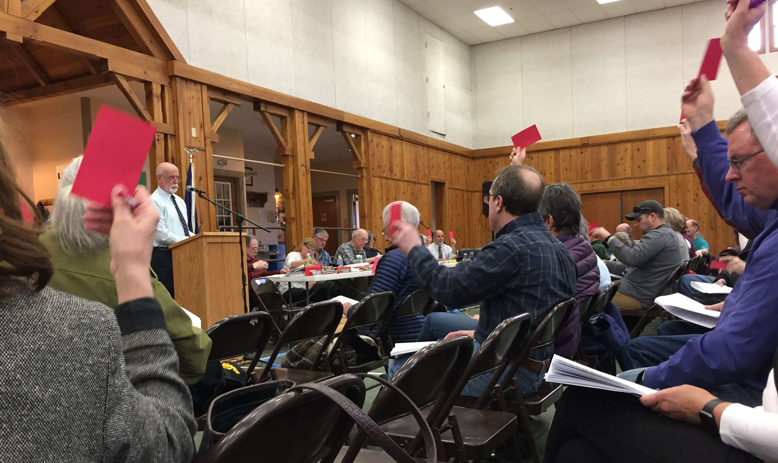 people sitting in chairs holding up red cards as a man at a lecturn speaks