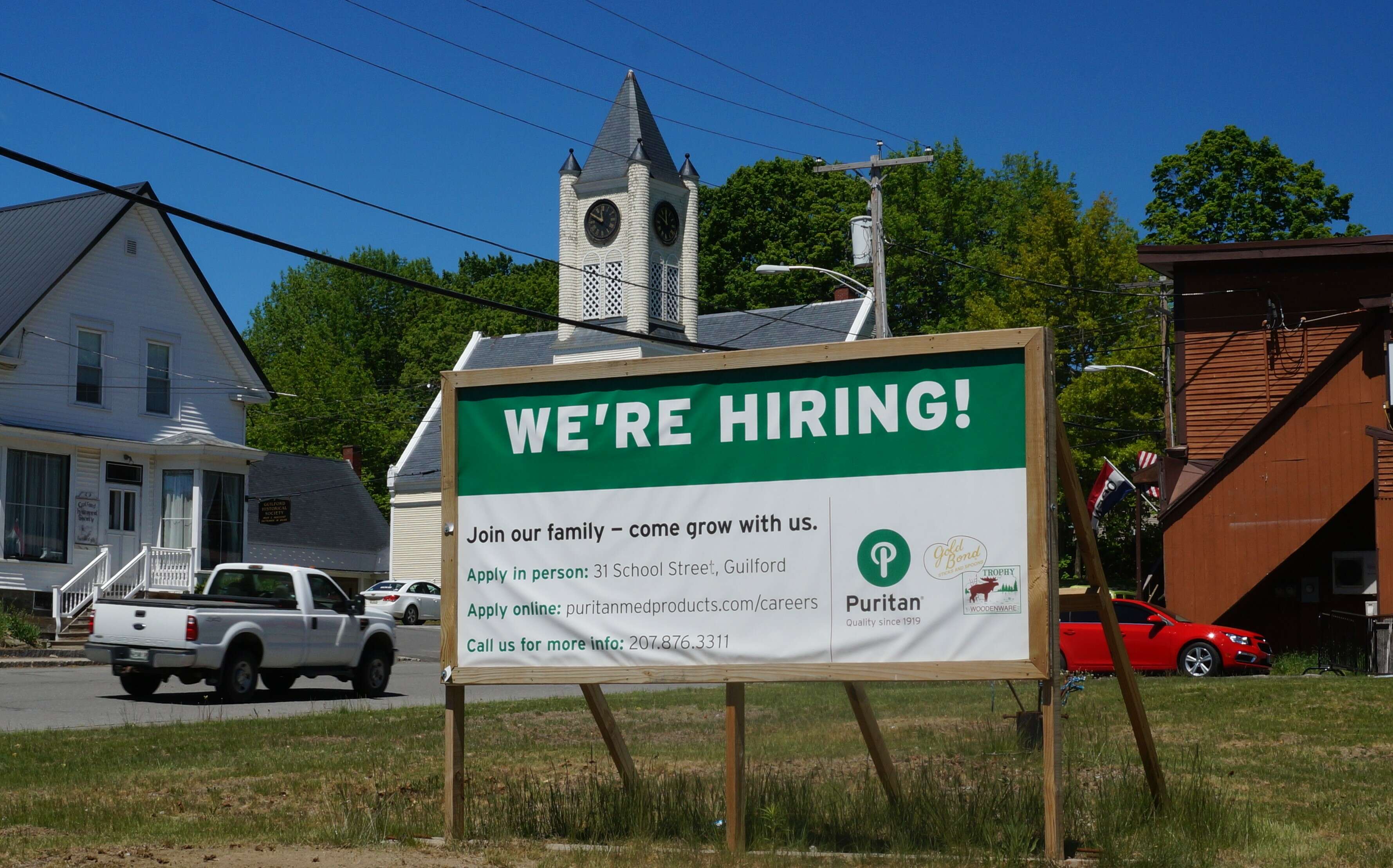 A sign that says we're hiring puritan medical products on a street with a wooden church and other small maine town buildings