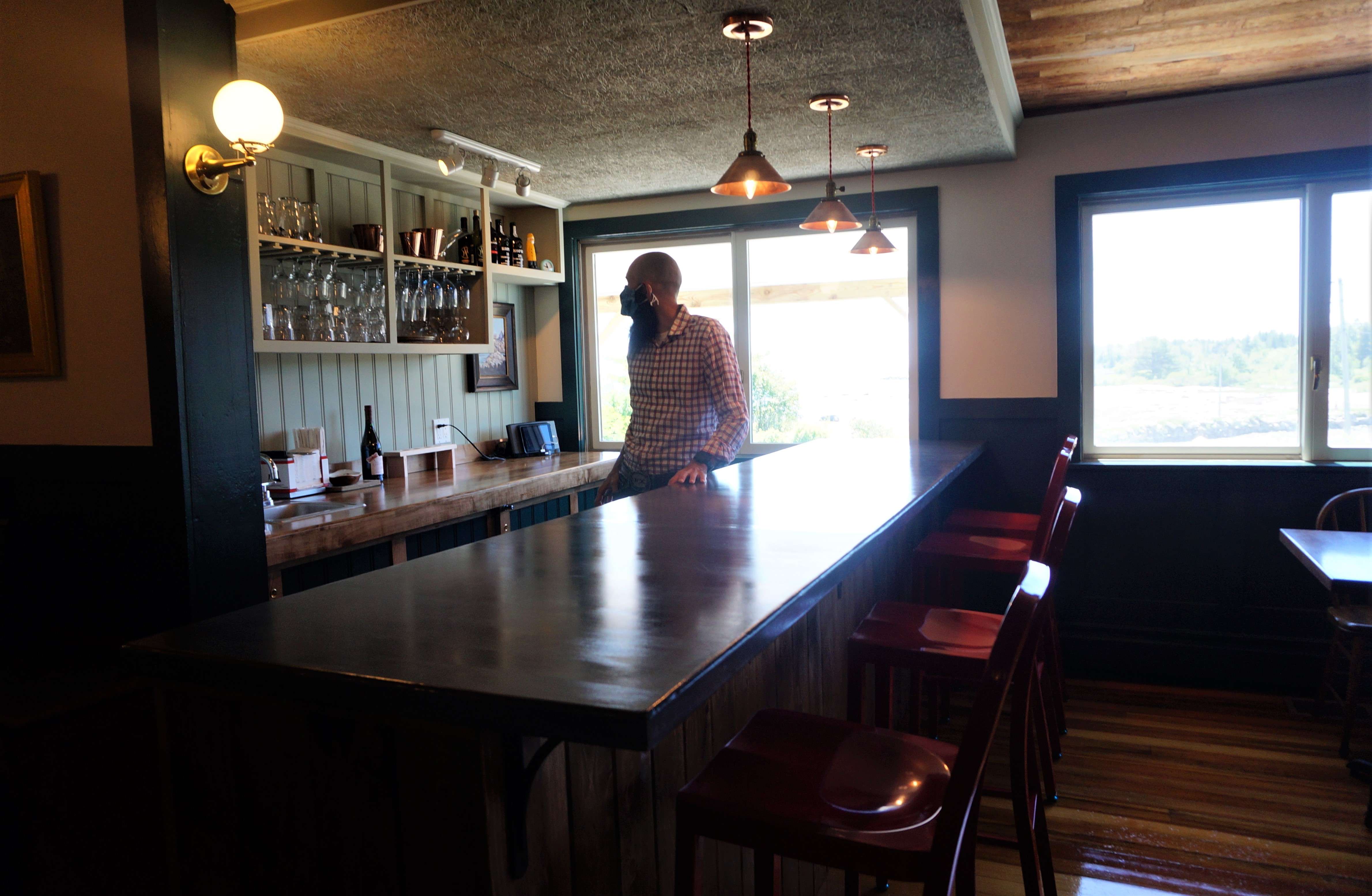 A man with a protective face covering stands behind a bar with a vew out the windows of the sea in the background