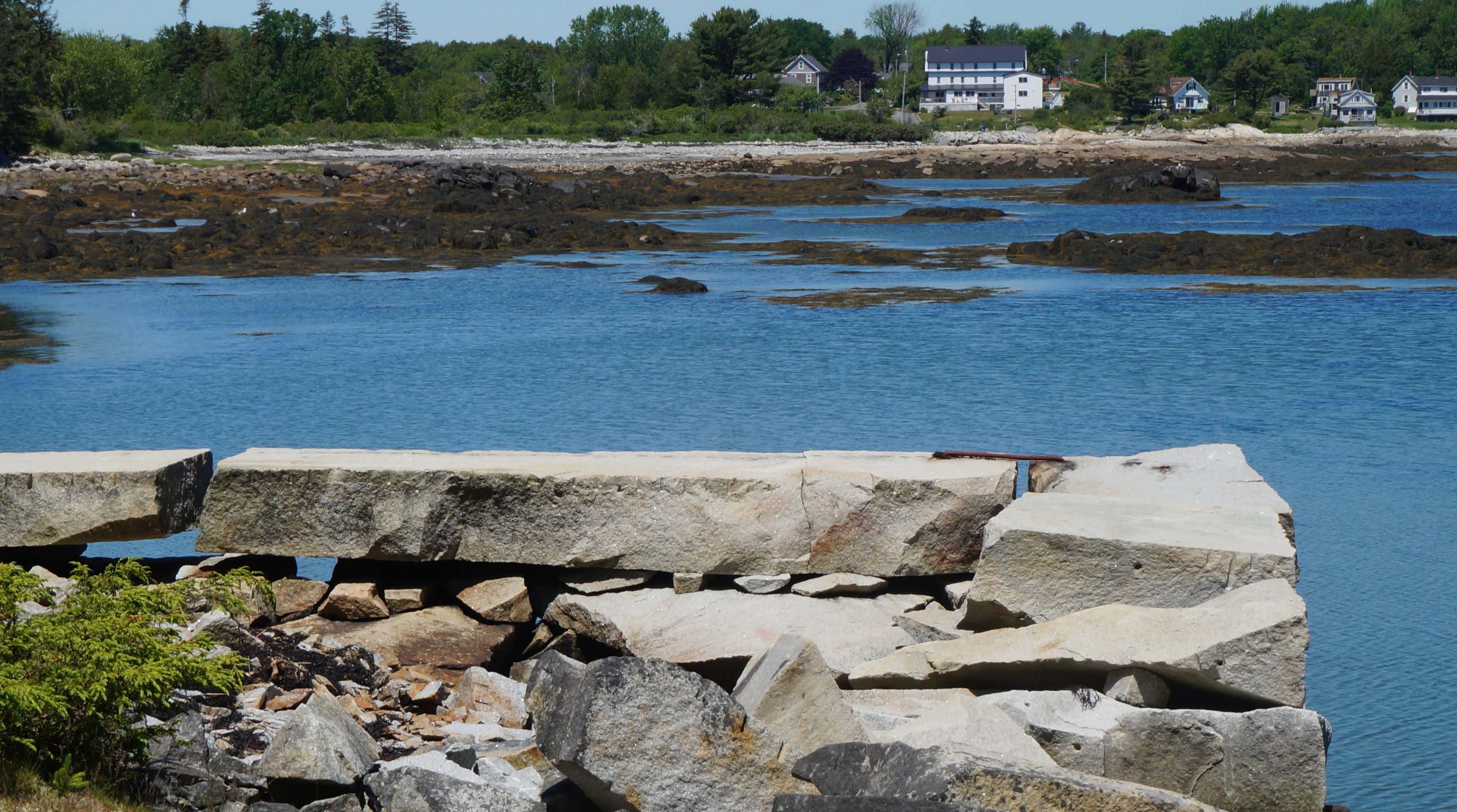 An old granite block pier in the foreground with deep blue ocean water and, beyond, a white three-story inn, the Craignair