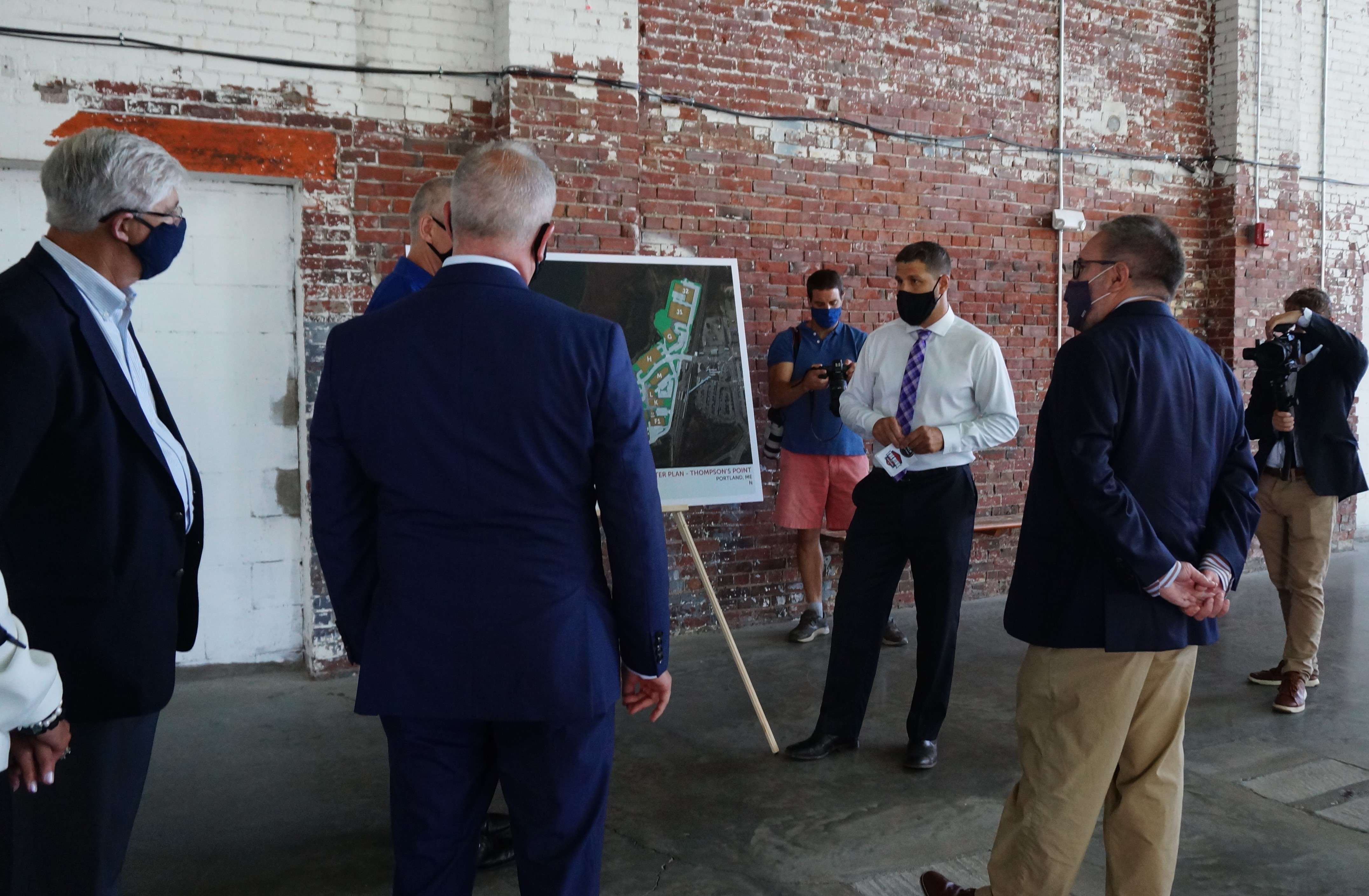 A man points to a diagram of a commercial development on an easel, as a bunch of men in suits look on.