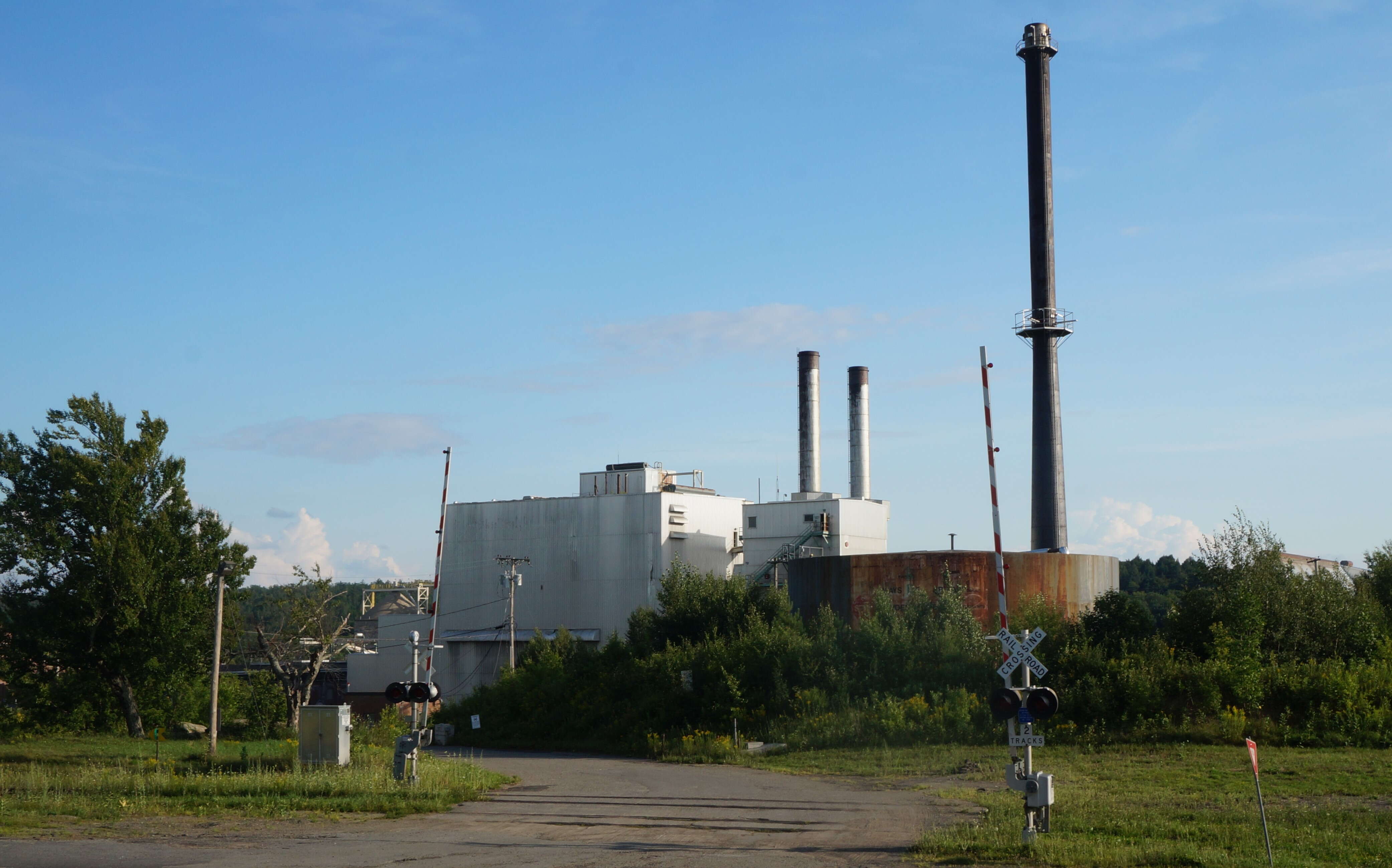 An old mill with a high smokestack stands behing weedy grass with a railroad crossing in the foreground