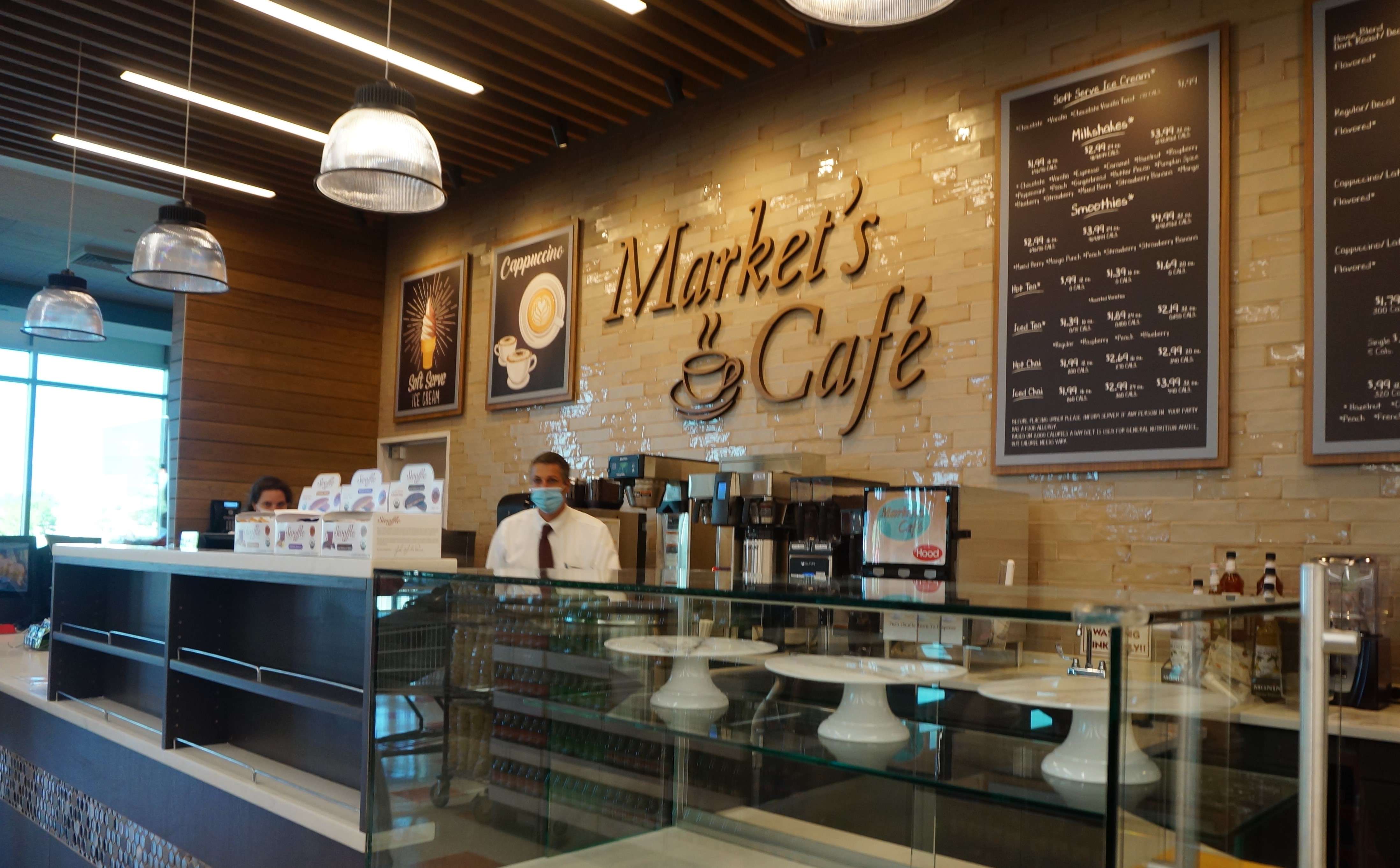 a man with a face covering stands behind a cafe counter next to a board advertising smoothies and coffee and baked goods