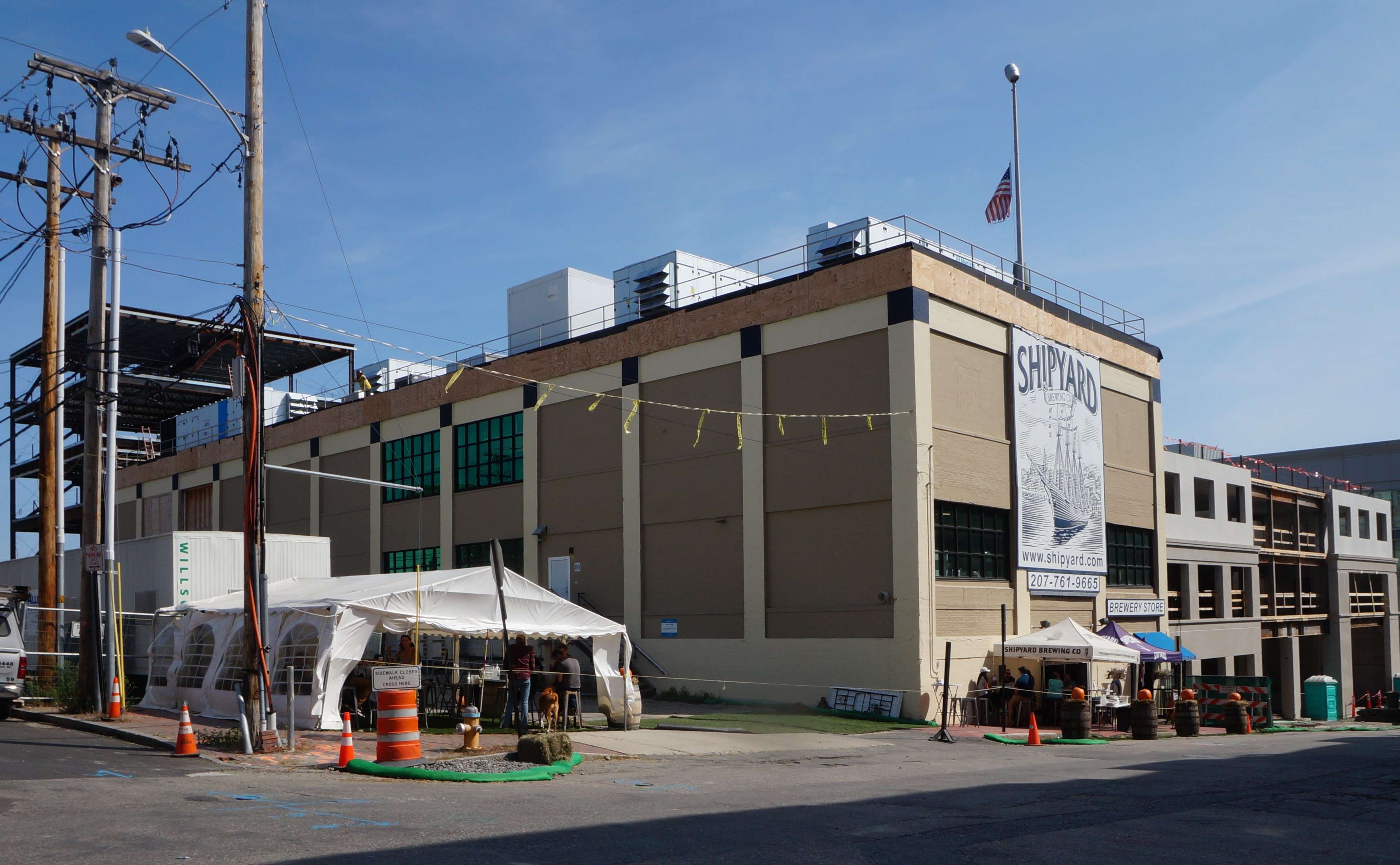 A tan three story cement building with a sign that says Shipyard Brewing Co with a modern building going up on its right and the steel structure of another new building behind it.