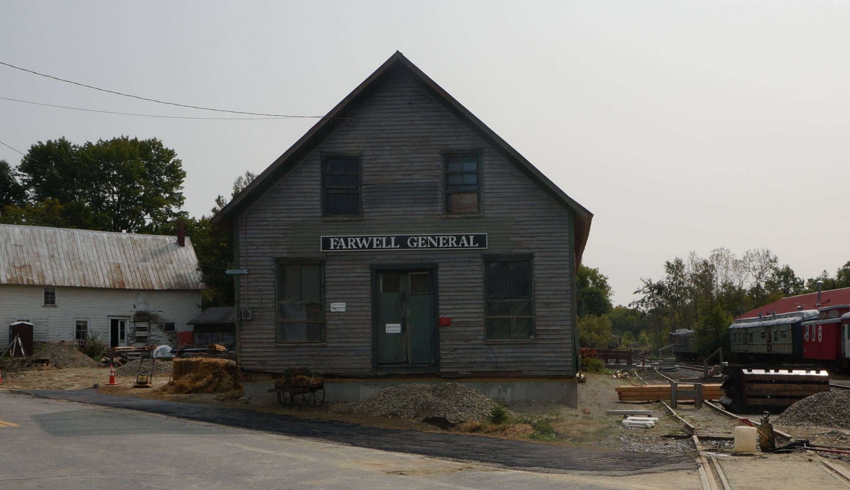 An old unpainted wooden building that says Farwell General on a sign above the door; to the left is an old white buiding with peeling paint and to the right railroad tracks with some box cars.