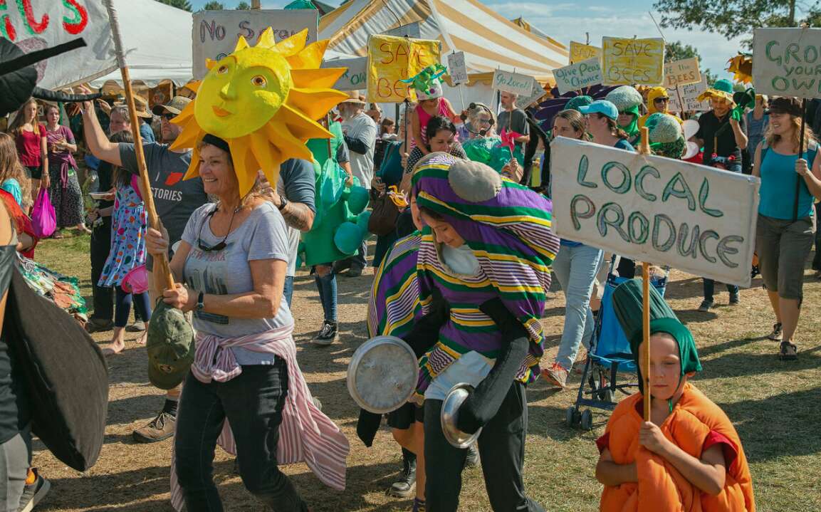 a group of people with signs that say things like local produce, including a small boy dressed as a carrot, march on a dirt road past vendor booths