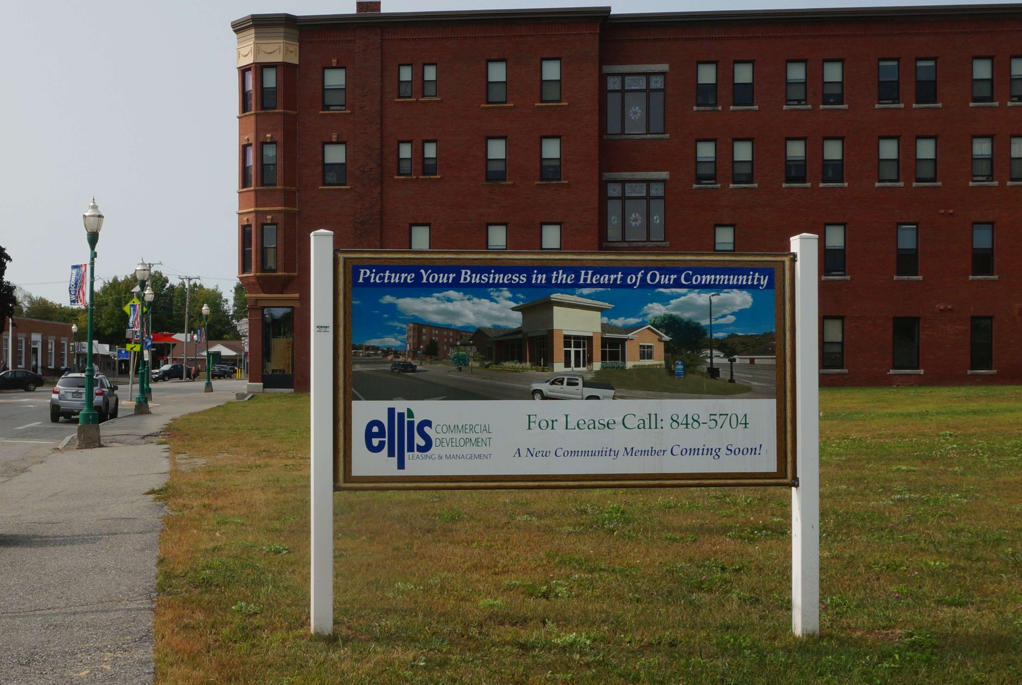 A sign in a grassy lot that says picture your business in this community with a four-story brick building in the background behind the lot