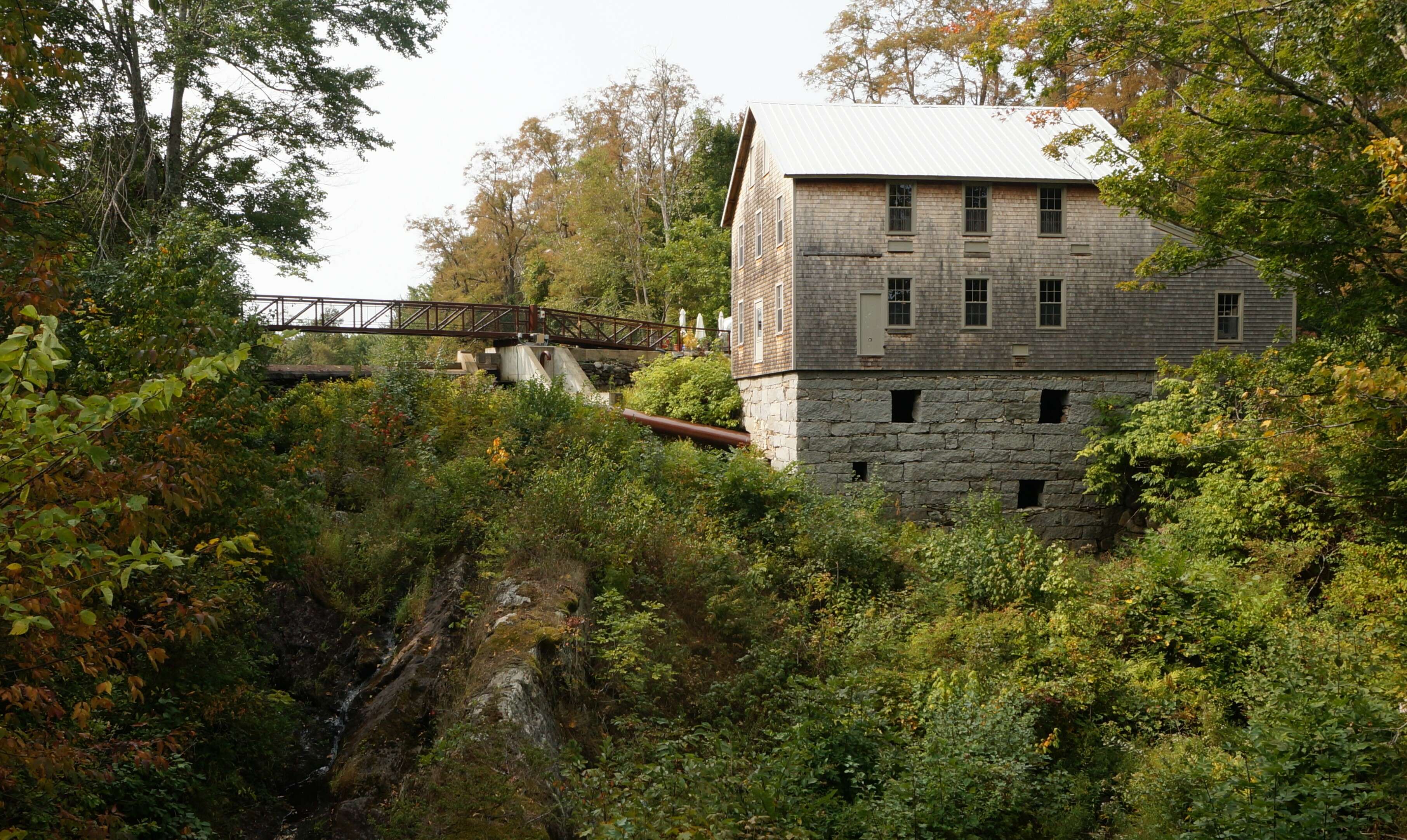 A mill with weathered shingles and a granite stone foundation on a vegatation rich ridge overlooking a rocky creek