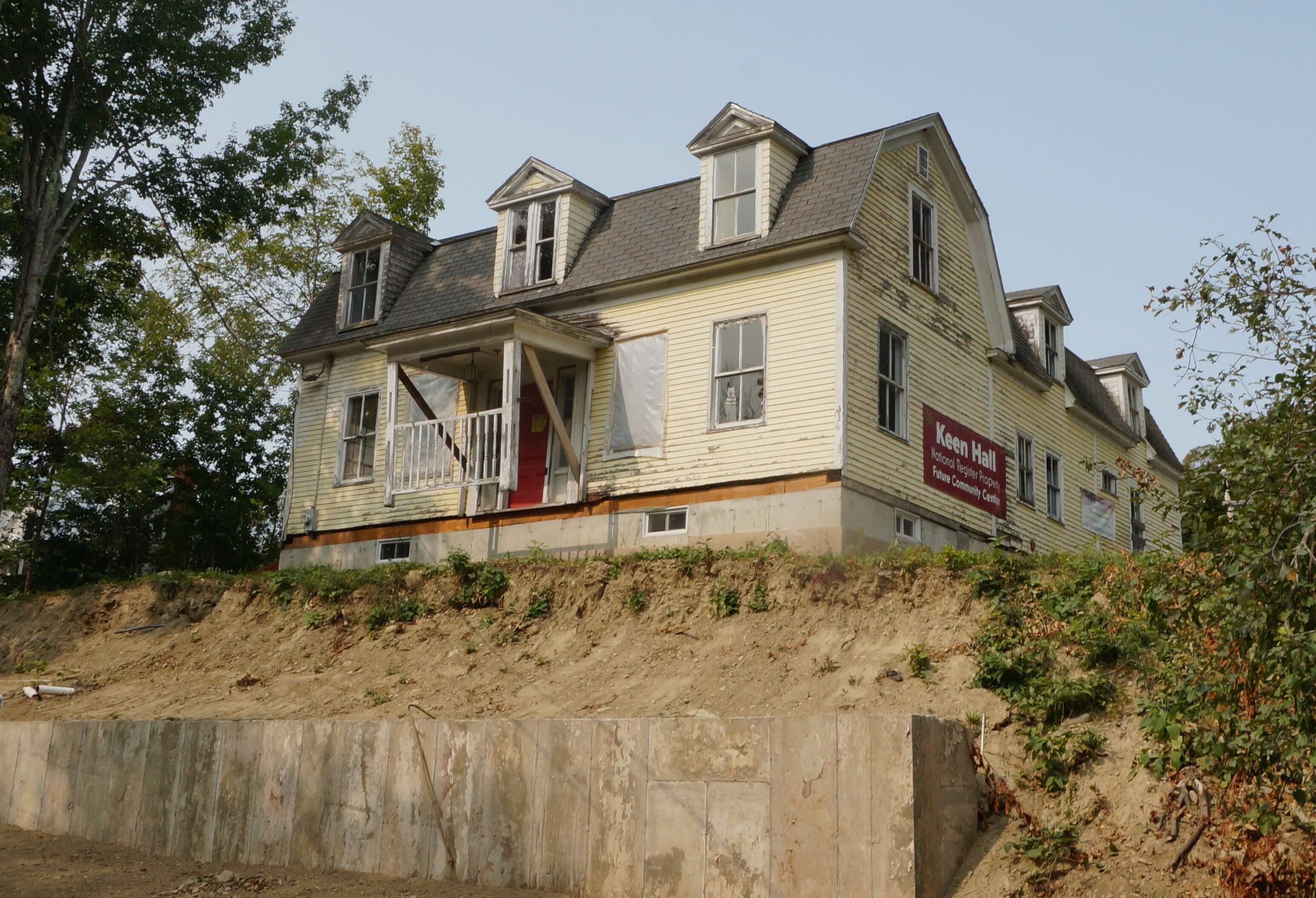 A two-century-old house with yellow peeling paint and dormers in a mansard roof sits atop a hill that has a new cement retaining wall at the bottom.