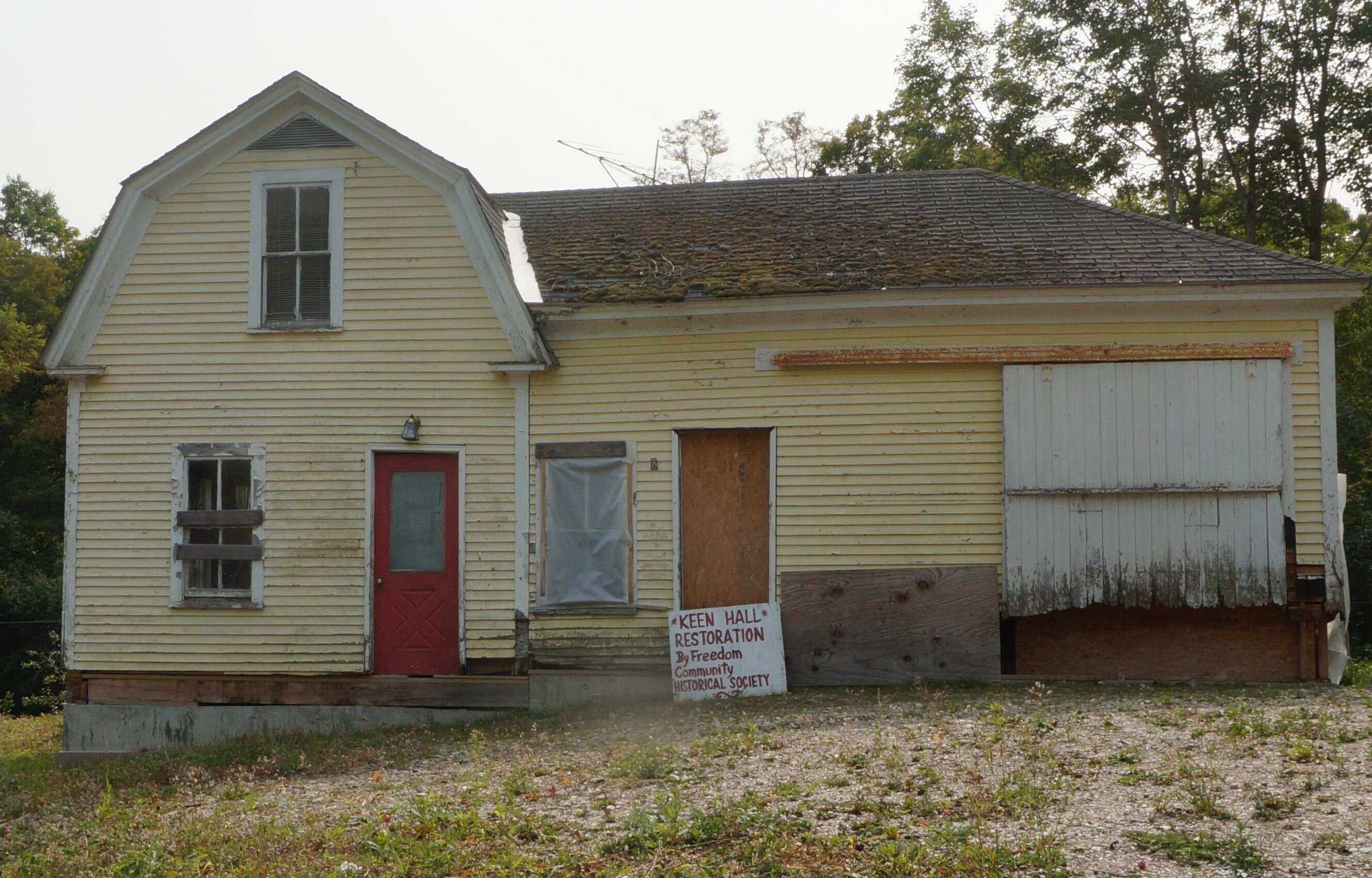 A rear view of an old house with peeling yellow paint and a hand-made sign leaning against it that says Keen Hall Freedom Community Historical Society