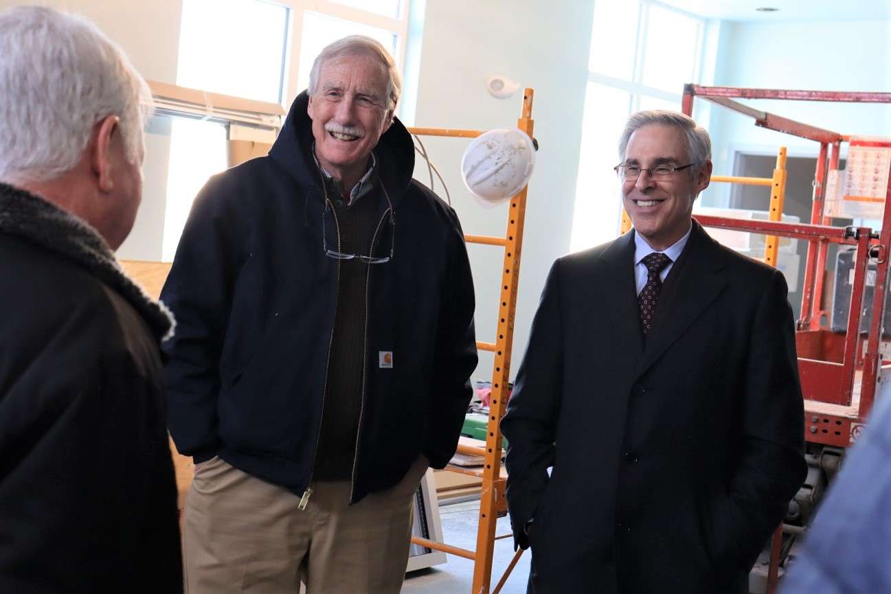 Two white-haired men talk to another white-haired man with his back to the camera. all three are wearing expensive winter coats at they're in an indoor construction site.