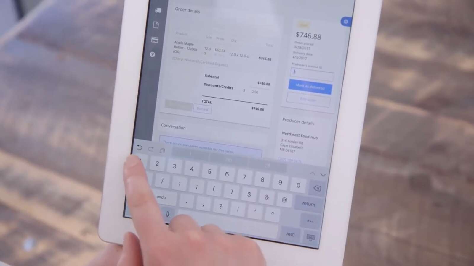 A finger pointing to a key on a digital tablet, with the screen on the tablet showing the price of apple butter and an order for it