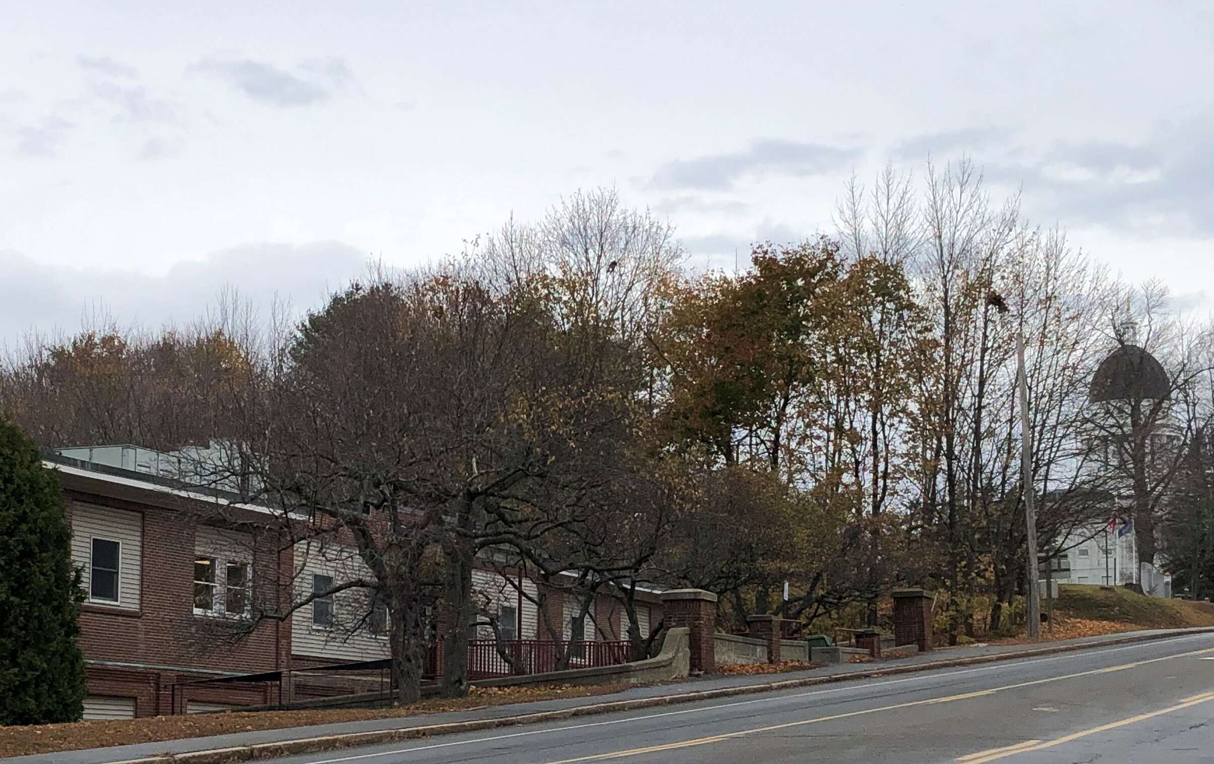 a low brick building on a gray fall day with a large granite building with a copper dome in the background, the Maine State House