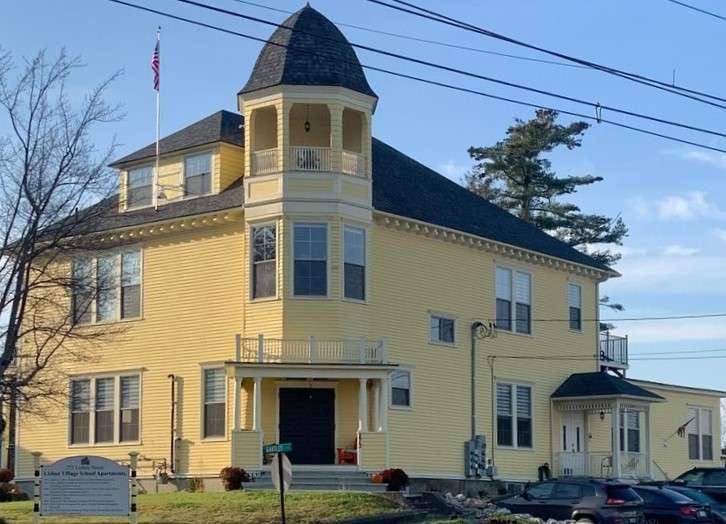 A yellow two-story wooden former school building with a cupola on the front and a sign that says lisbon village school apartments