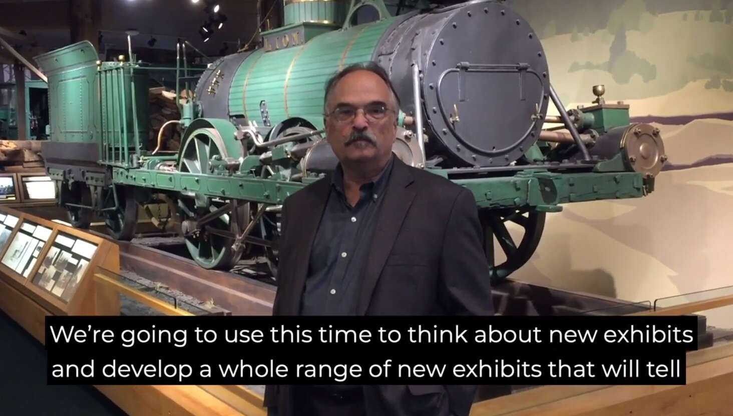 A man standing in front of a display of an old train engine and words on the screen that say we're going to use this time to think about new echibits and develop and wholr range of new exhibits
