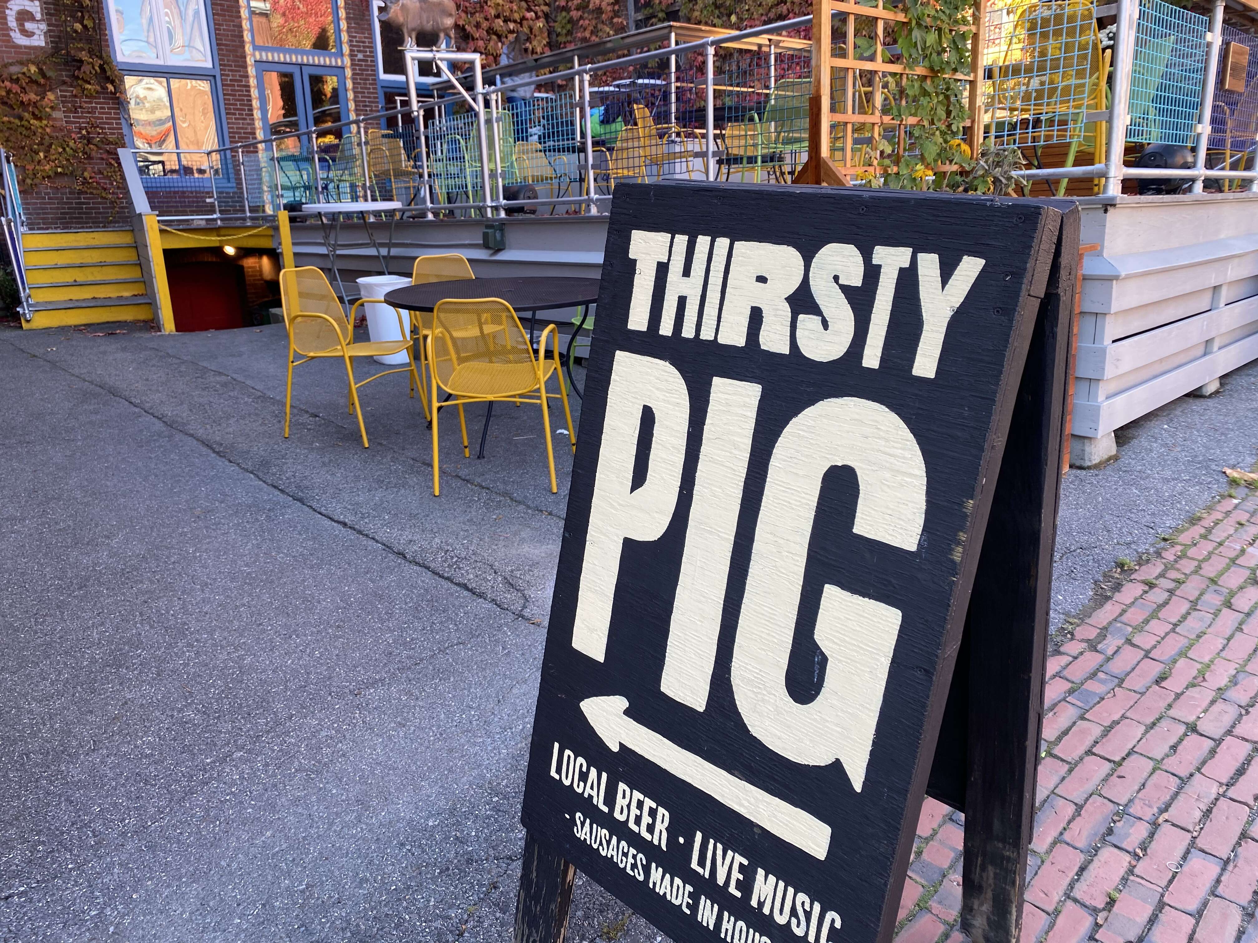 A sidewalk sign that says Thirsty Pig, live music, beer, etc, in front of deck with chairs and tables