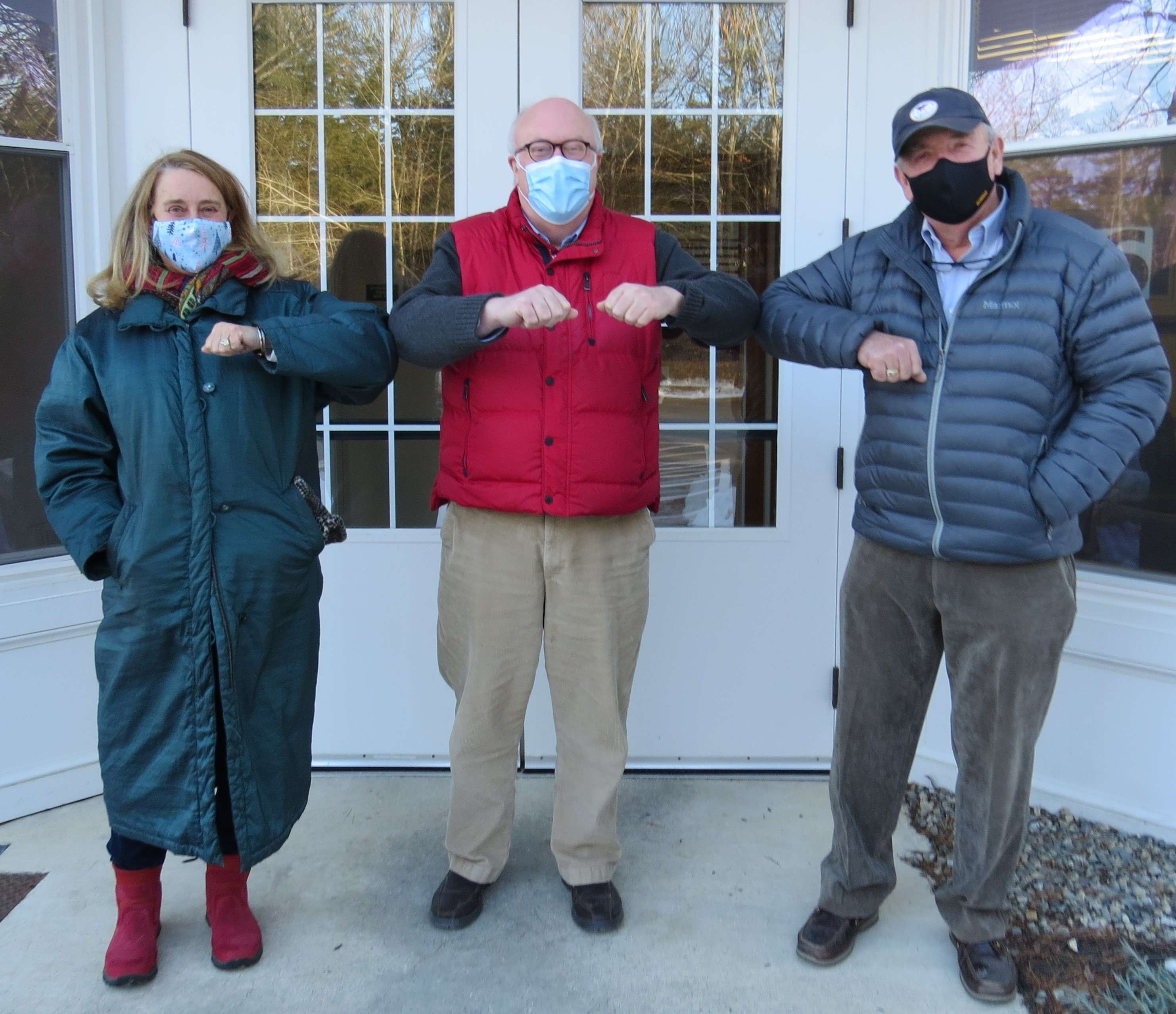 A woman and two men stand in front of a door wearing face coverings and winter clothing doing elbow bumps for the camera