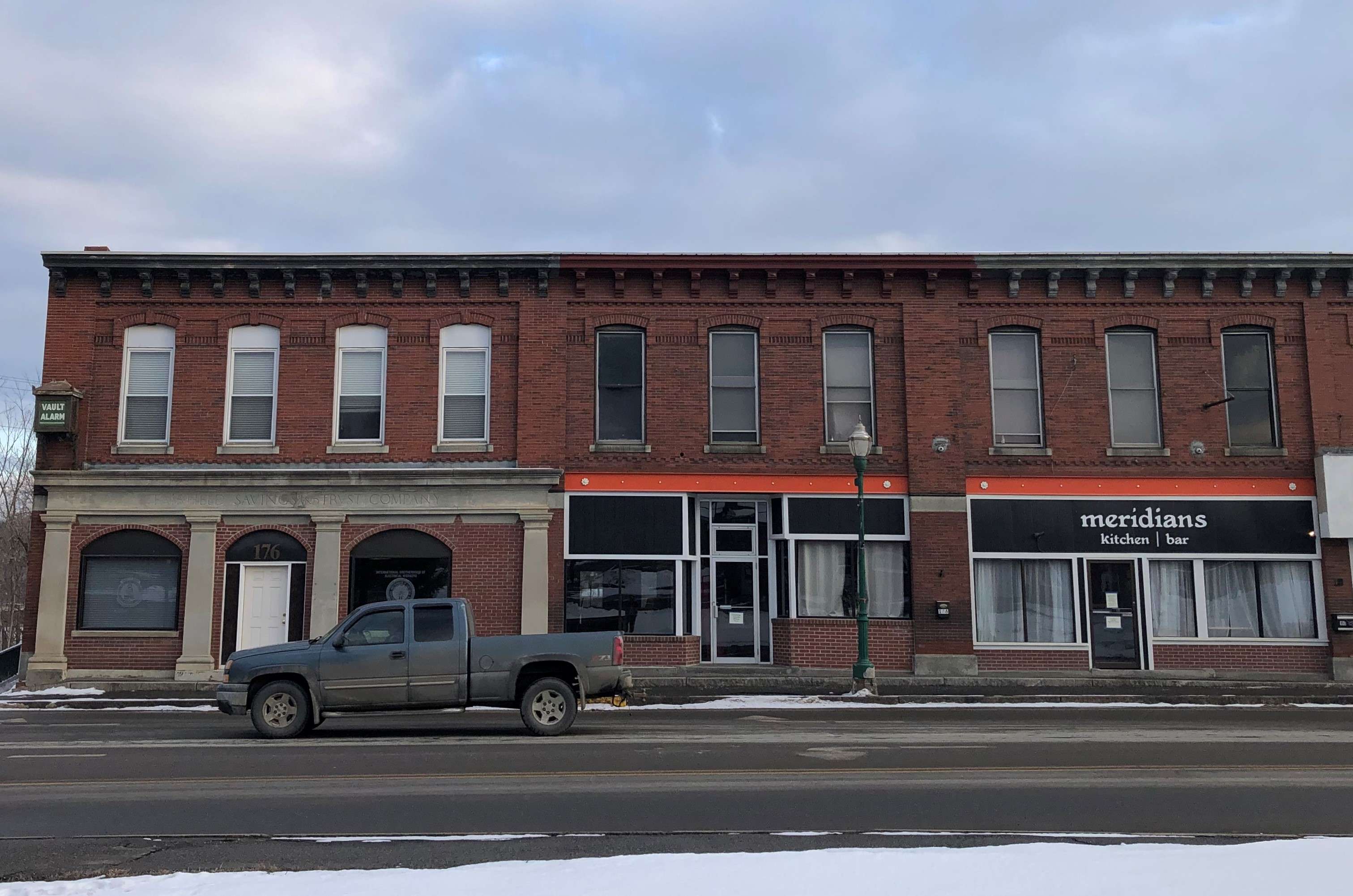 A row of 19th century two-story brick buildings with granite details, one has a black and orance sign that says meridian, a pickup truck goes by on the street