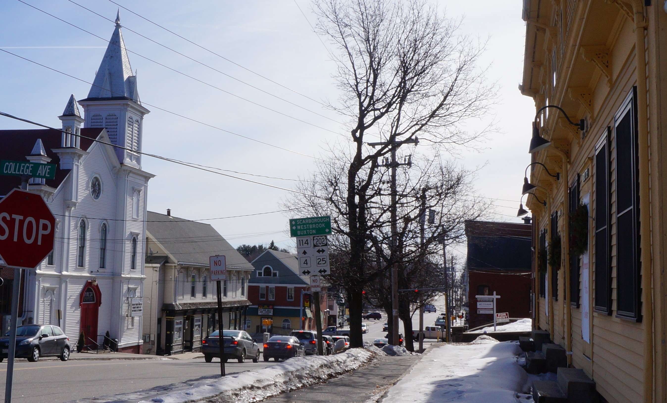 A main street with a white steepled church older clapboard and brick buildings melting dirty snow and a line of cars at a red light at an intersection that shows signs for routes 114, 202, 4 and 25