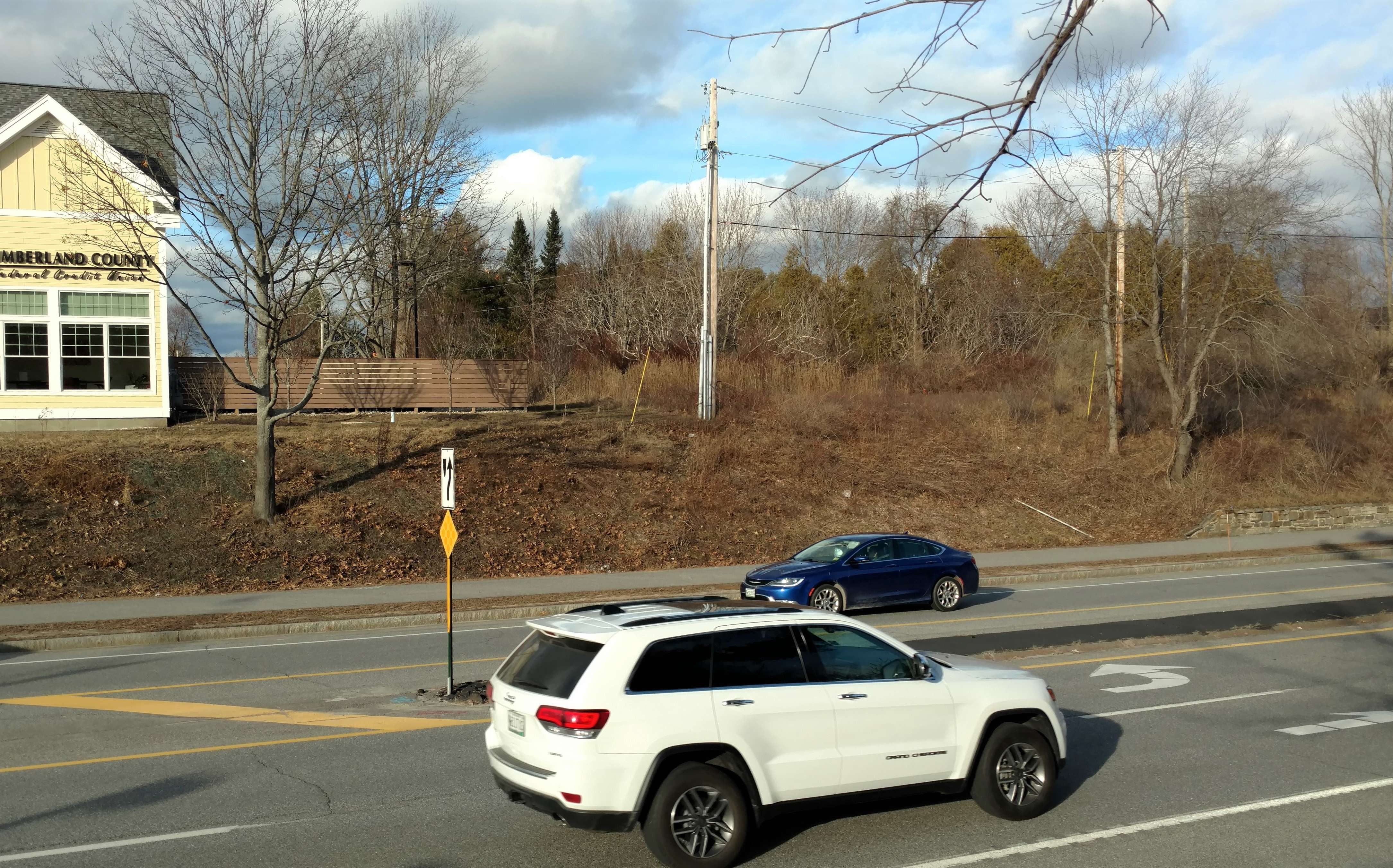 yellow gabled building to the left of vacant, overgrown lot, with cars passing in front