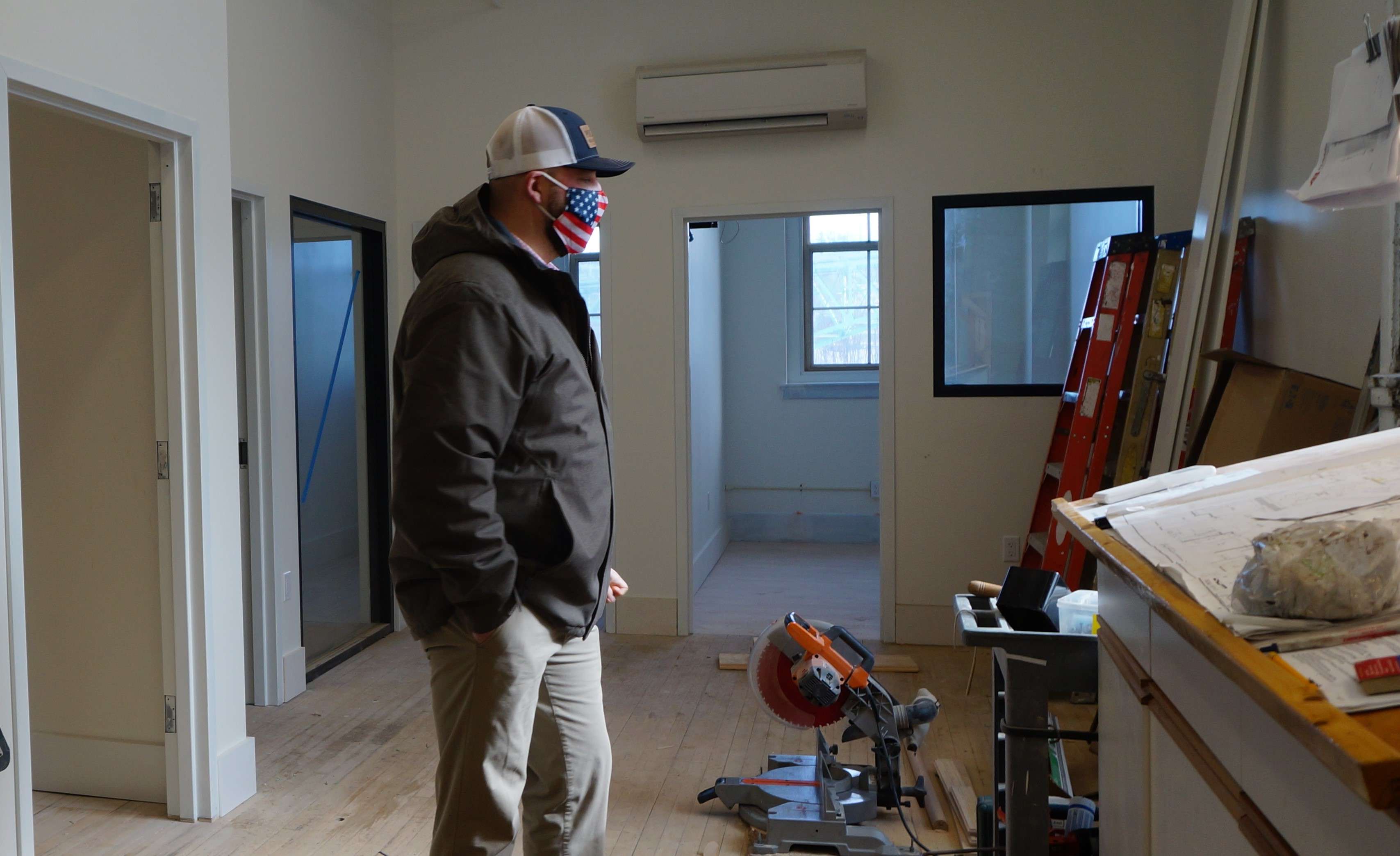 a man wearing an american flag mask stands in sunny space under construction on a wooden floor looking ou tthe window