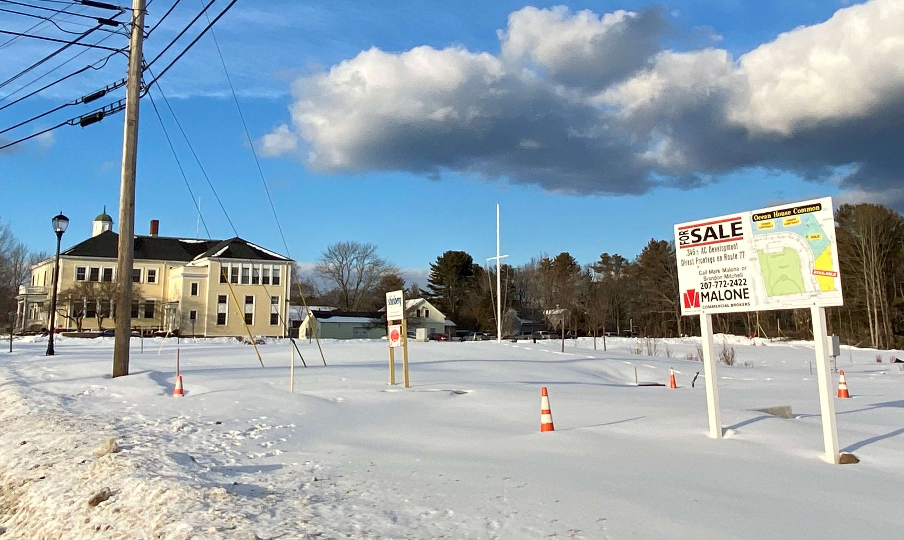 a snowy expanse with a real estate broker's sign showing lots with a large clapboard yellow building, cape elizabeth's town hall, in the background