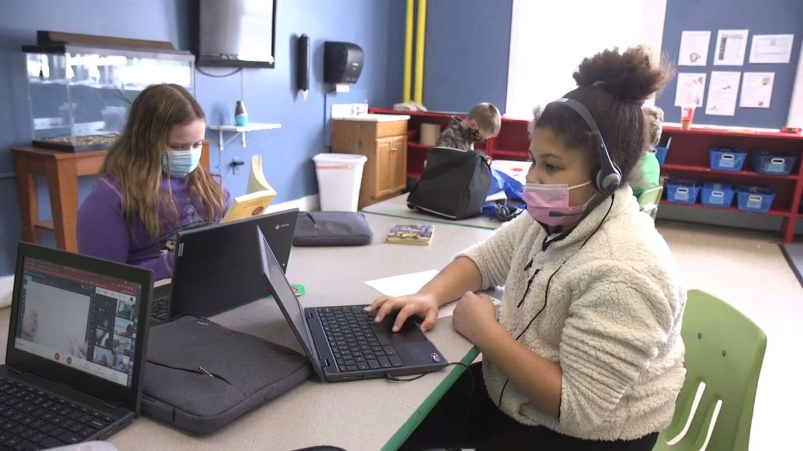 Two young girls, one white and one black, work on laptops at a table in a large room will other kids take part in activities in the background