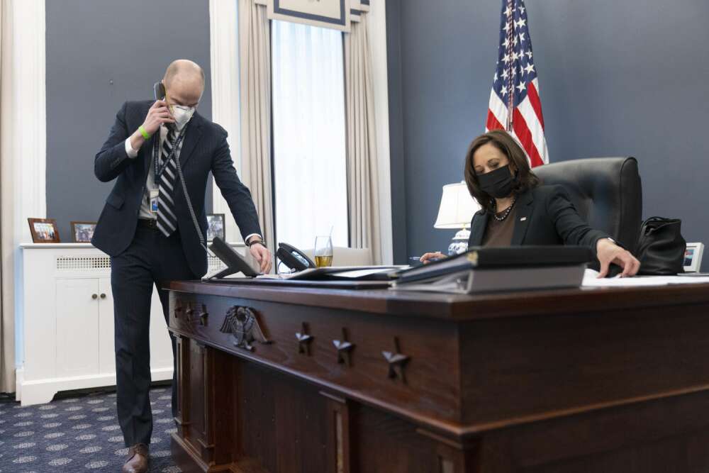  vice president Kamala Harris, a Black woman wearing a face mask, sits behind a large ornate wooden desk working on papers; a white man in a face covering is on a telephone standing up next to the desk