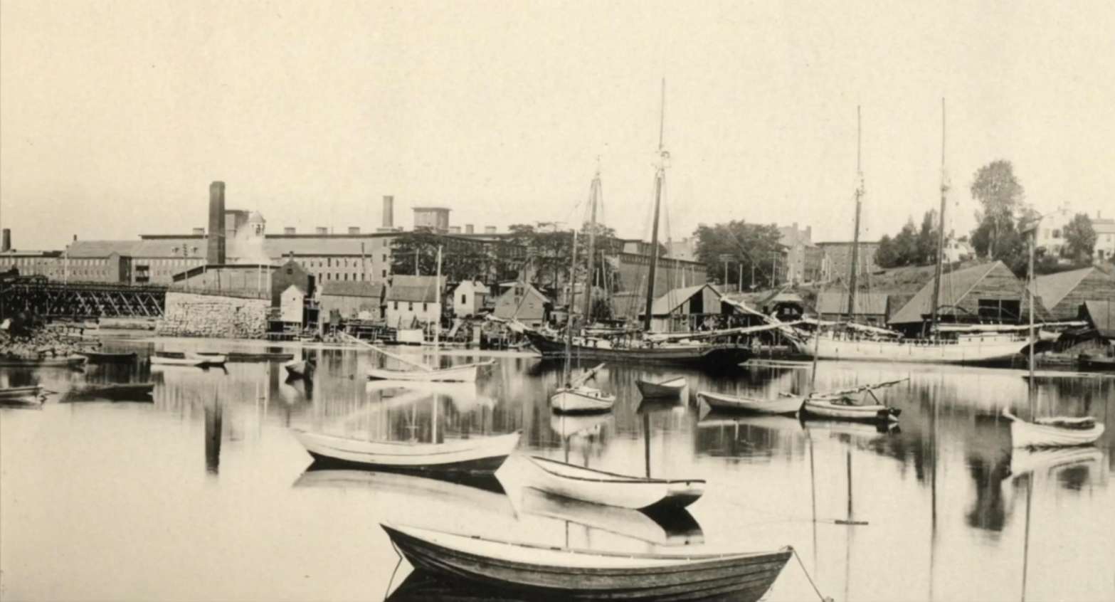 a sepia toned photo from likely the early 20th century of boats in a river with land with buildings on it behind