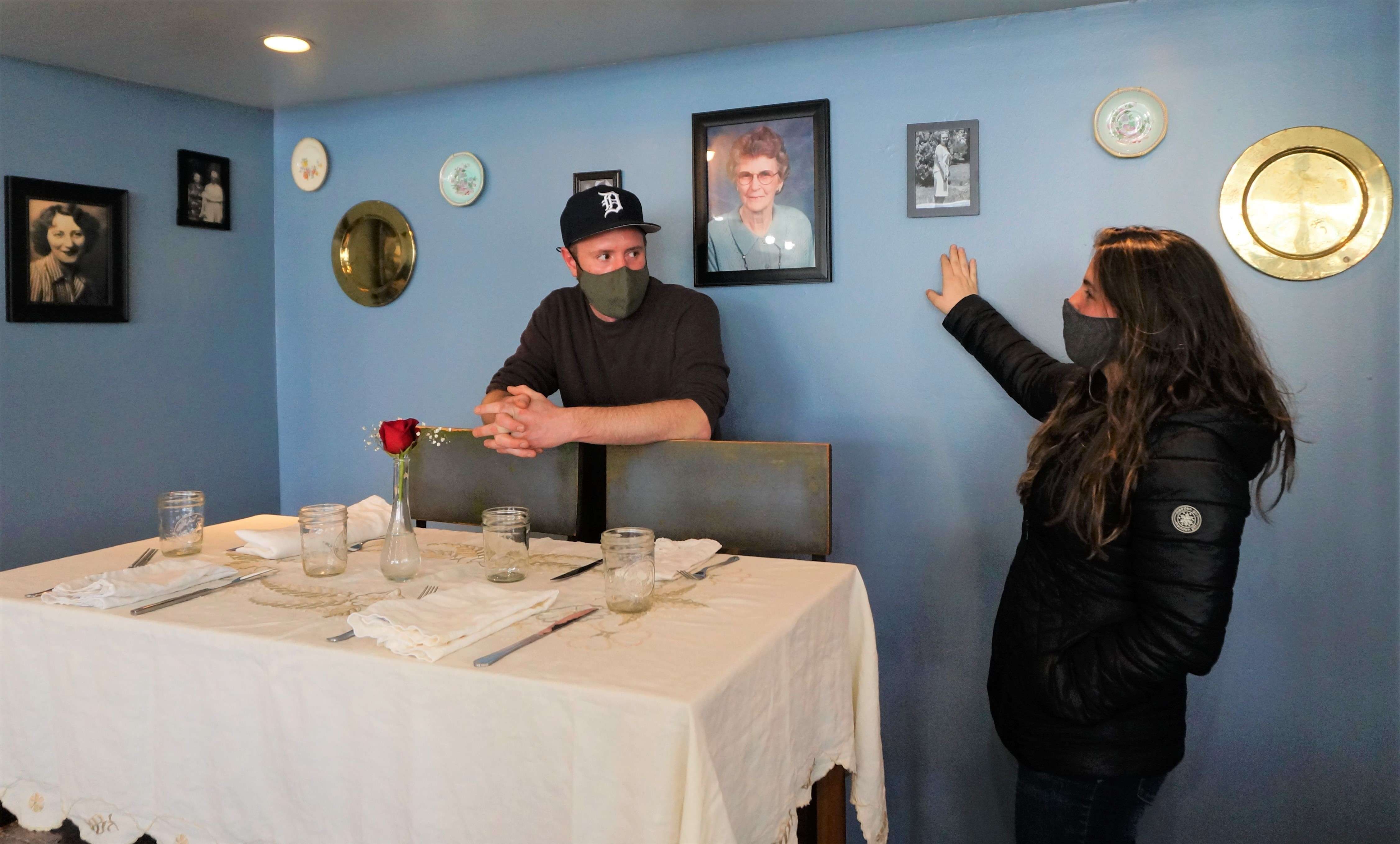 A white man with a mask and a Detroit Tigers hat leans on a chair behind a dining table set with a white table cloth and napkins, while a woman points to a photo of an older woman on the wall.