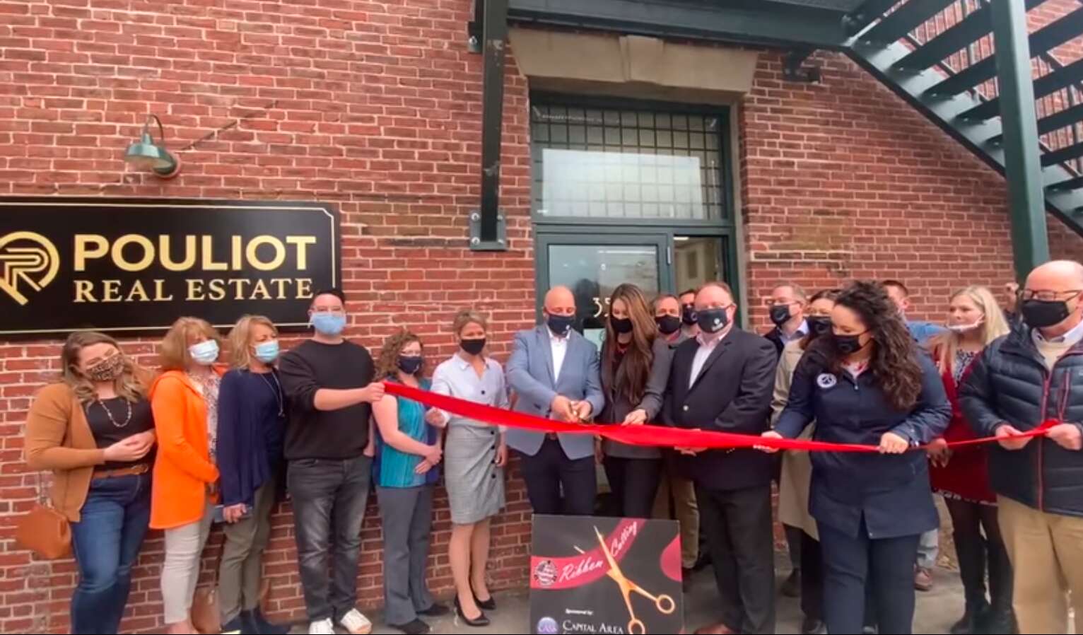 a man, white in a gray suit, cuts a thick red ribbon with giant scissors as a group of poeple stands around him behind them is a brick wall with a sign that says pouliot real estate