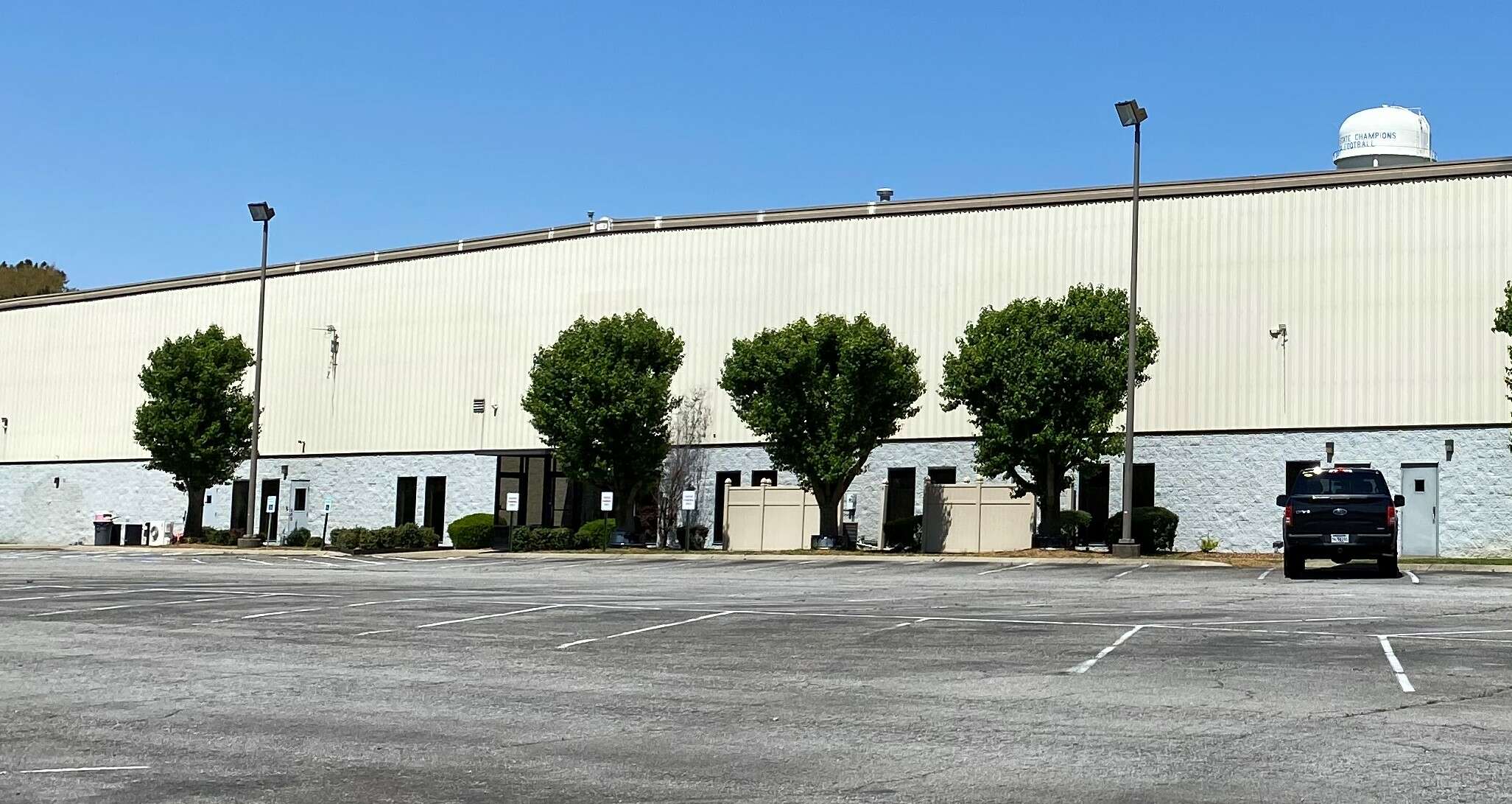 A very large manufacturing building with a faux stone lower level painted white and corrugated metal upper level, some doors but no windows, and trees along the walls