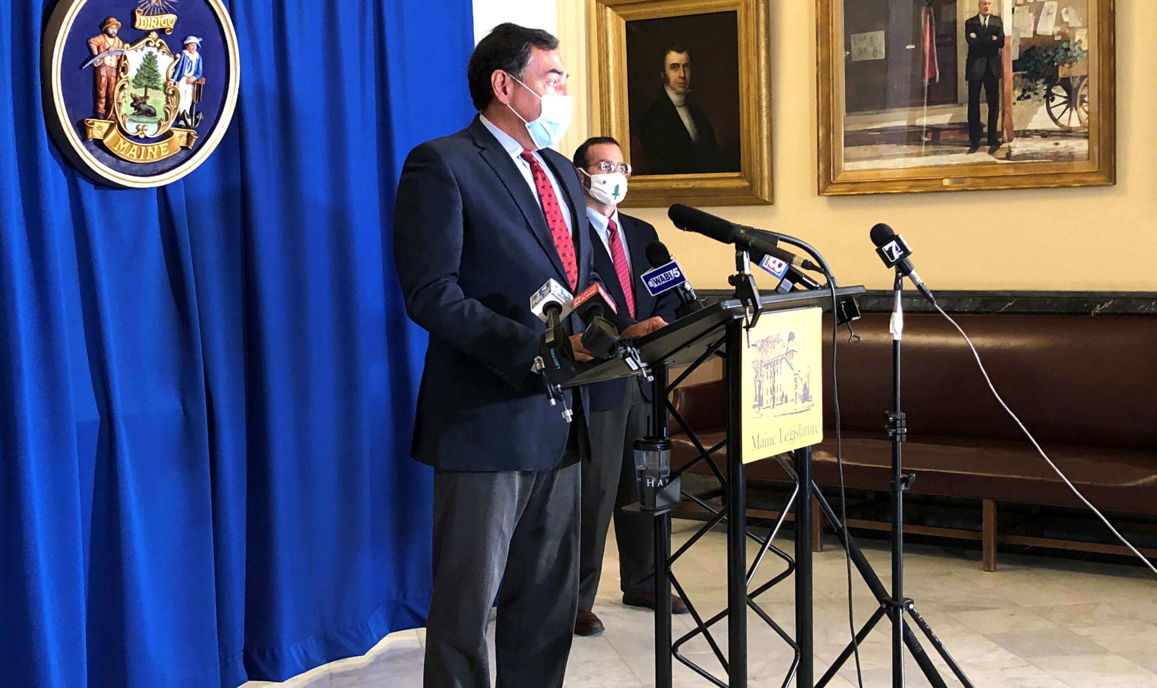 A man, white, wearing a face covering and in a suit, stands at a lecturn with microphones on it as another man stands in the background. there is a blue backdrop behind them with the seal of the state of maine on it, and portions of painted portraits can be seen on the walls.