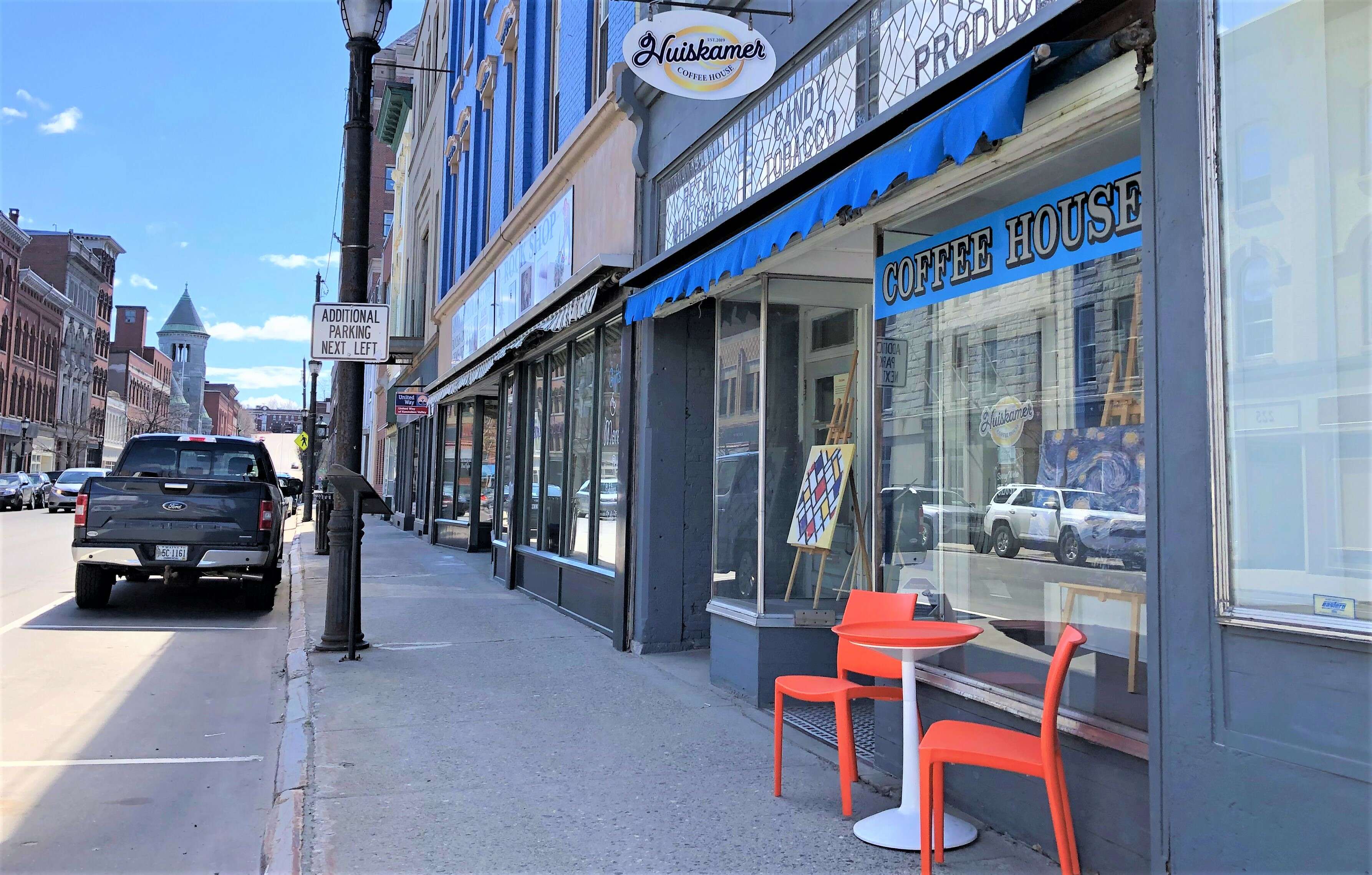 A bright downtown storefront has a small organge table and two chairs on the narrow sidewalk in the front the sign says Huiskamer Coffee House.