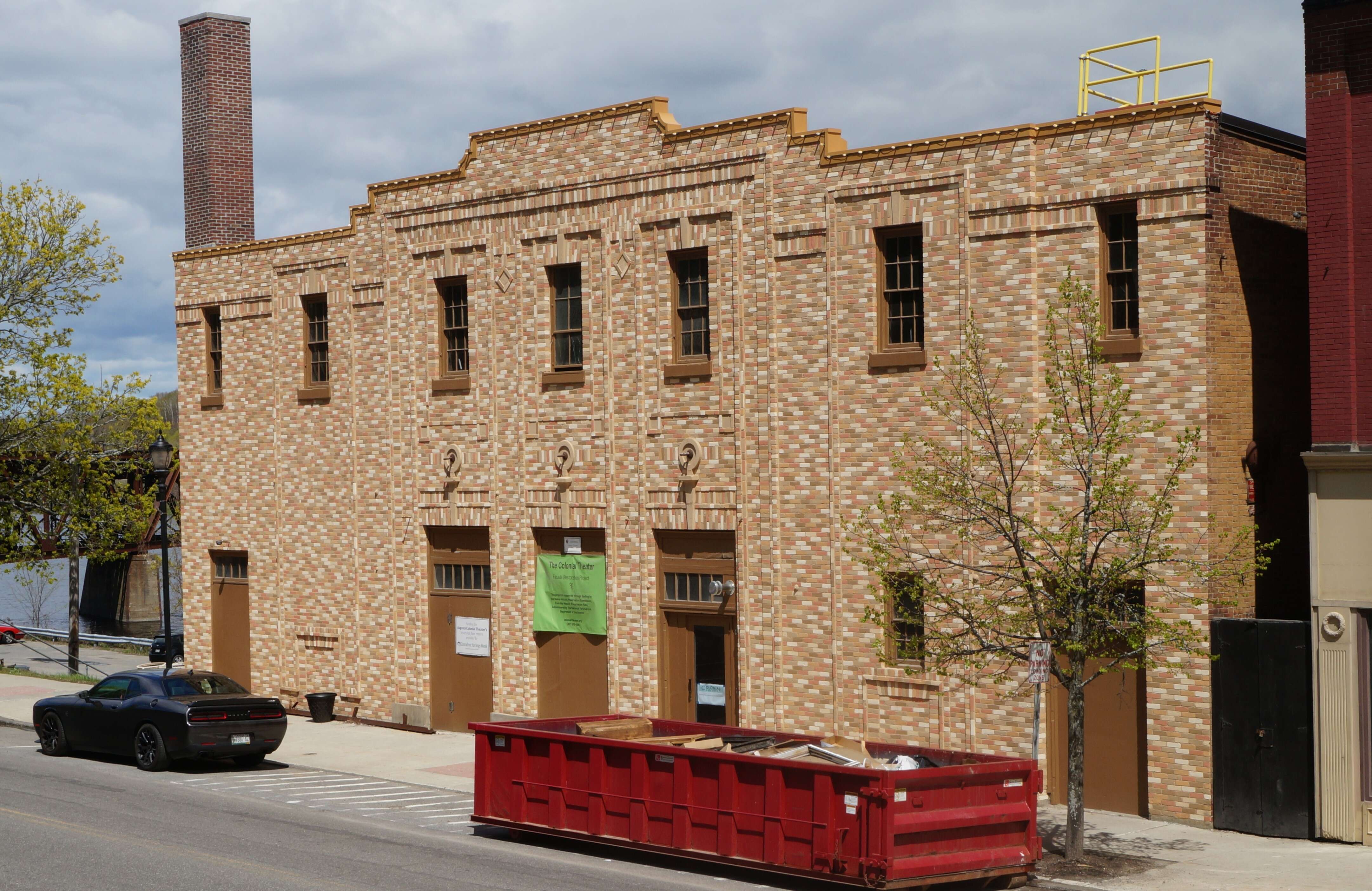 A yellow brick theater building in art deco style with a large red dumpster in front and a glimpse of a river behind it.