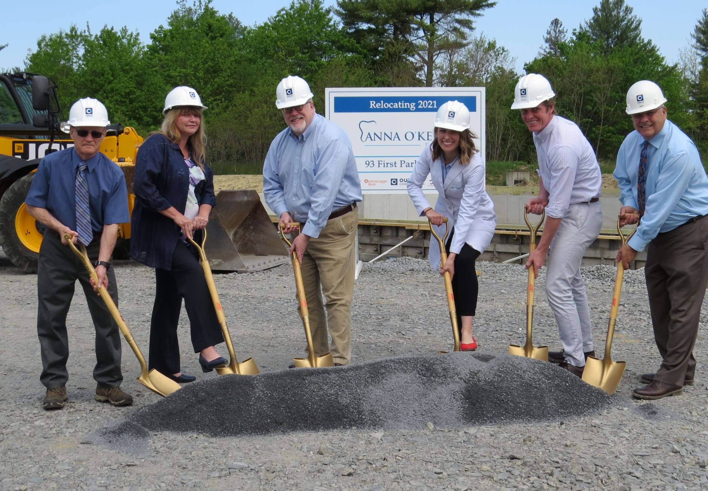 a group of six people, four men and three women, white, wearing hardhats, pose with shovels in a pile of granite