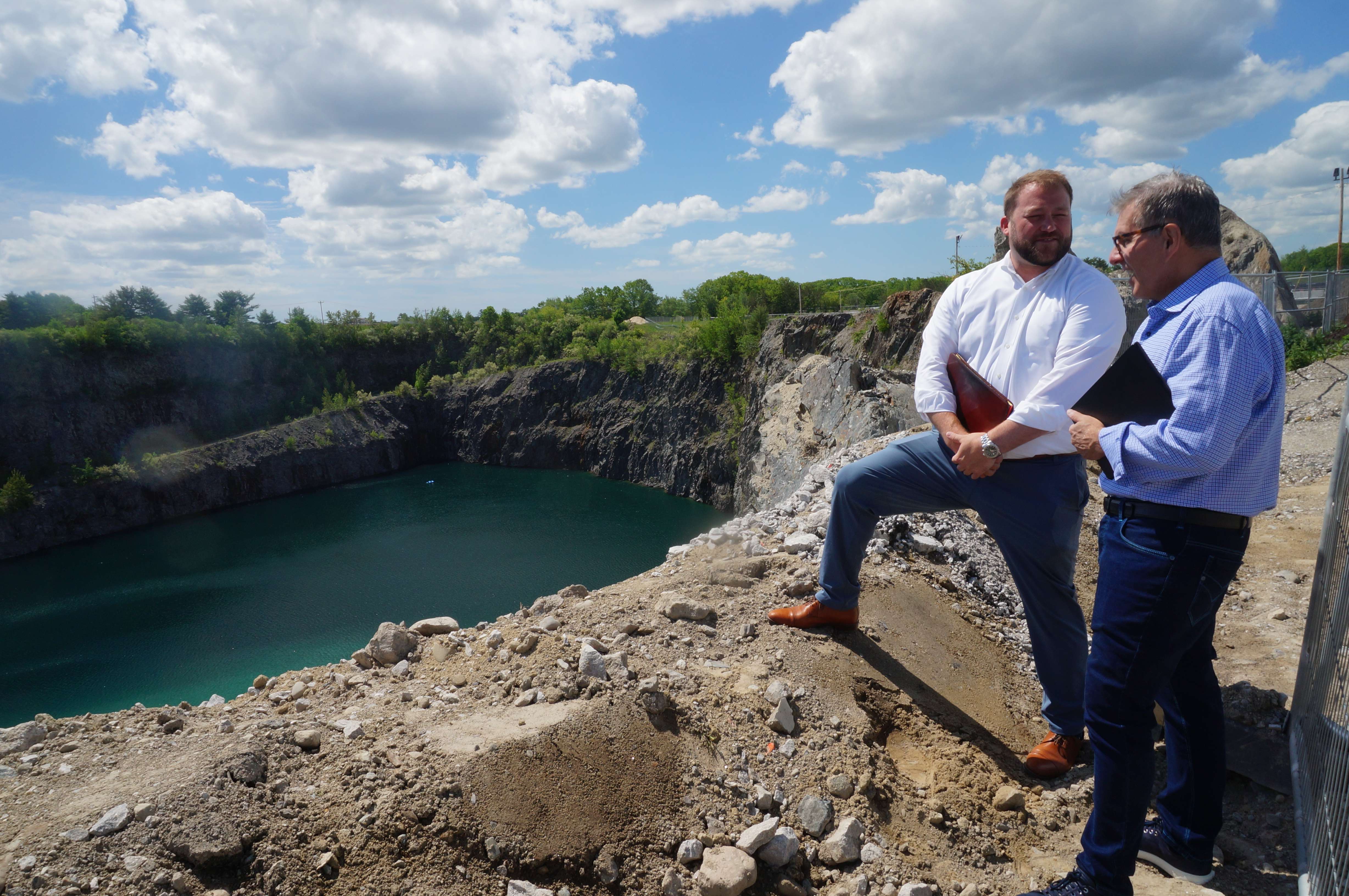 two men, both white, one with a beard, stand on the edge of a deep quarry with brigh green water at the bottom