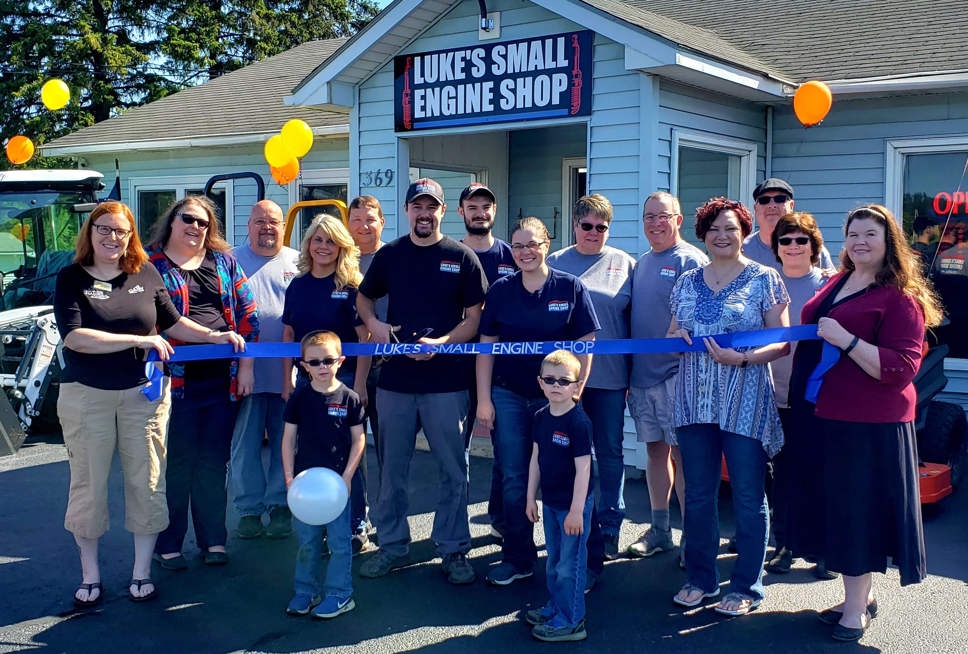 A group of people stands in front of a ceremonial ribbon as the man in the middle, holding a giant pair of scissors smiles at the camera. A sign on the building behind them says Lukes Small Engine Shop.