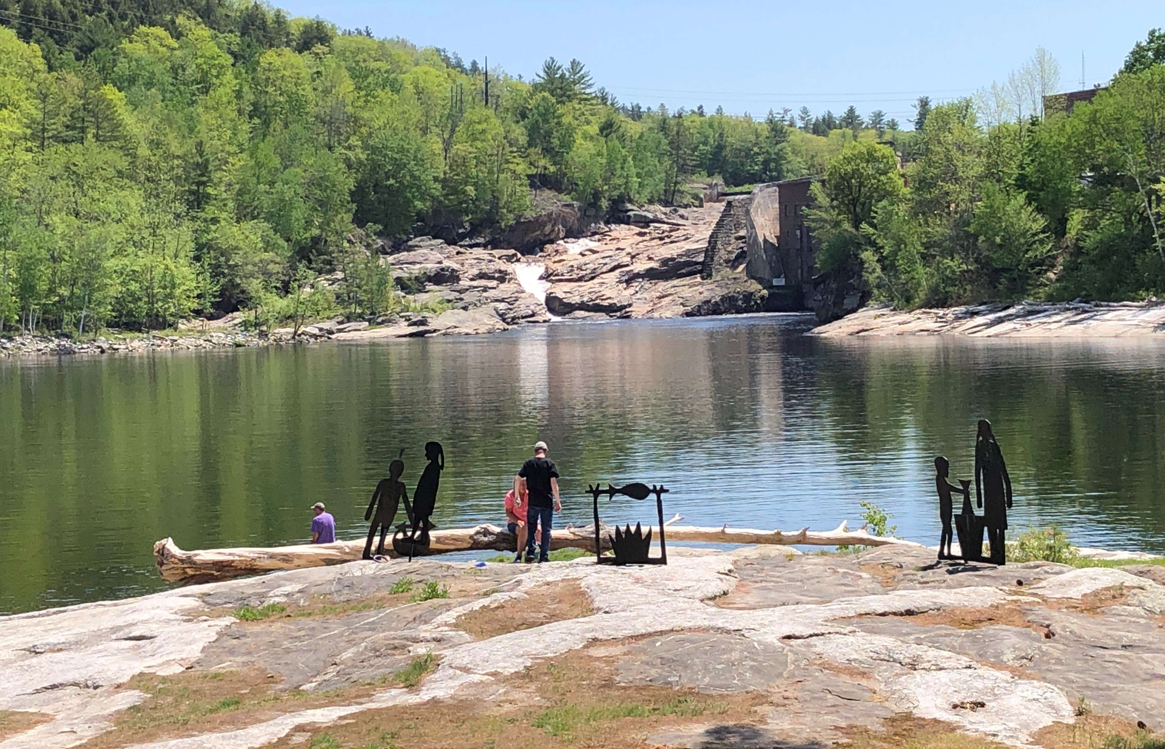 people and public art that looks like people stand on the rocks overlooking the androscoggin river and rocky rumford falls