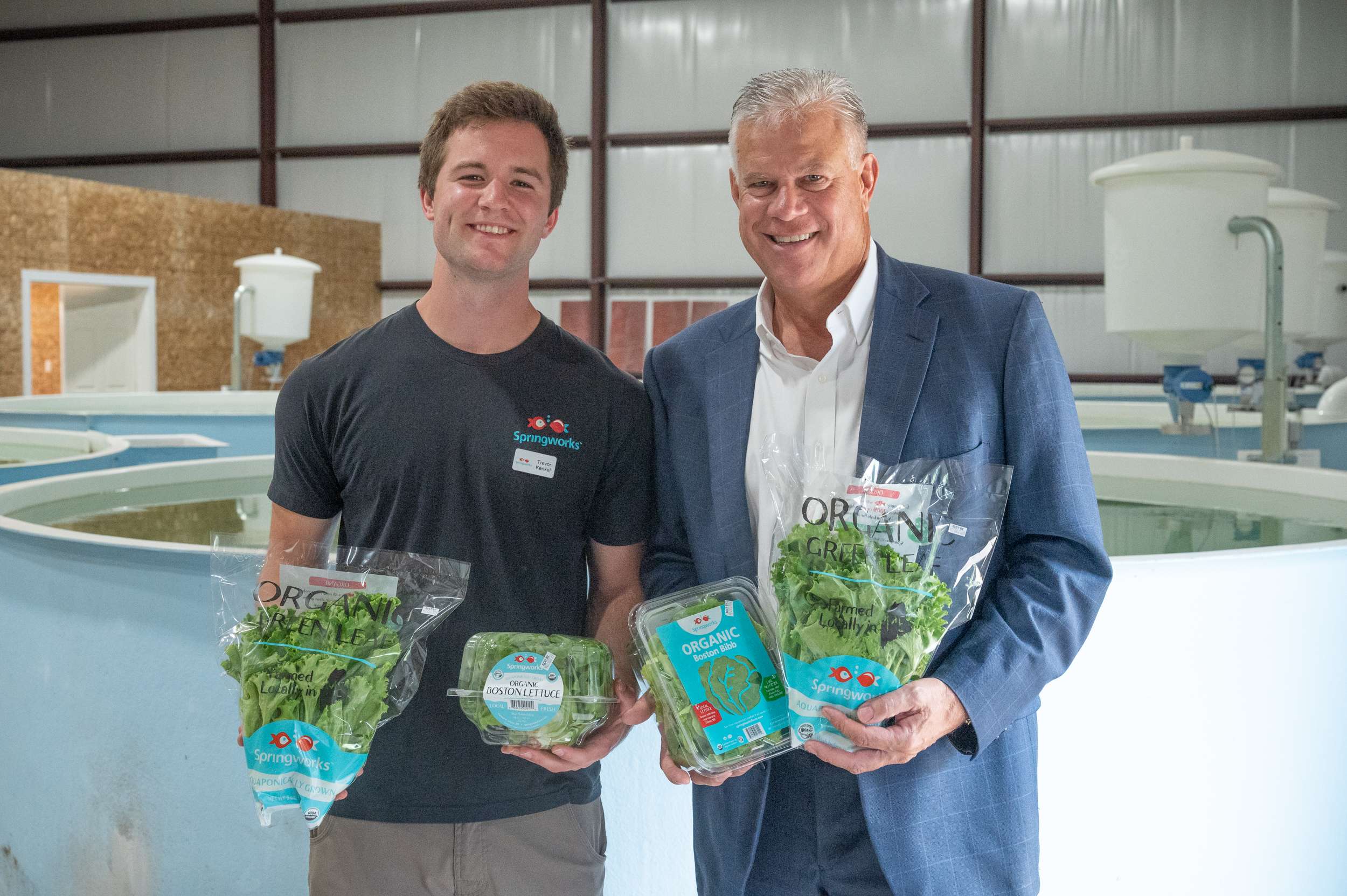 A young man holds two bags of lettuce that say Springworks Farm on them next to an older man, also holding two bags of lettuce