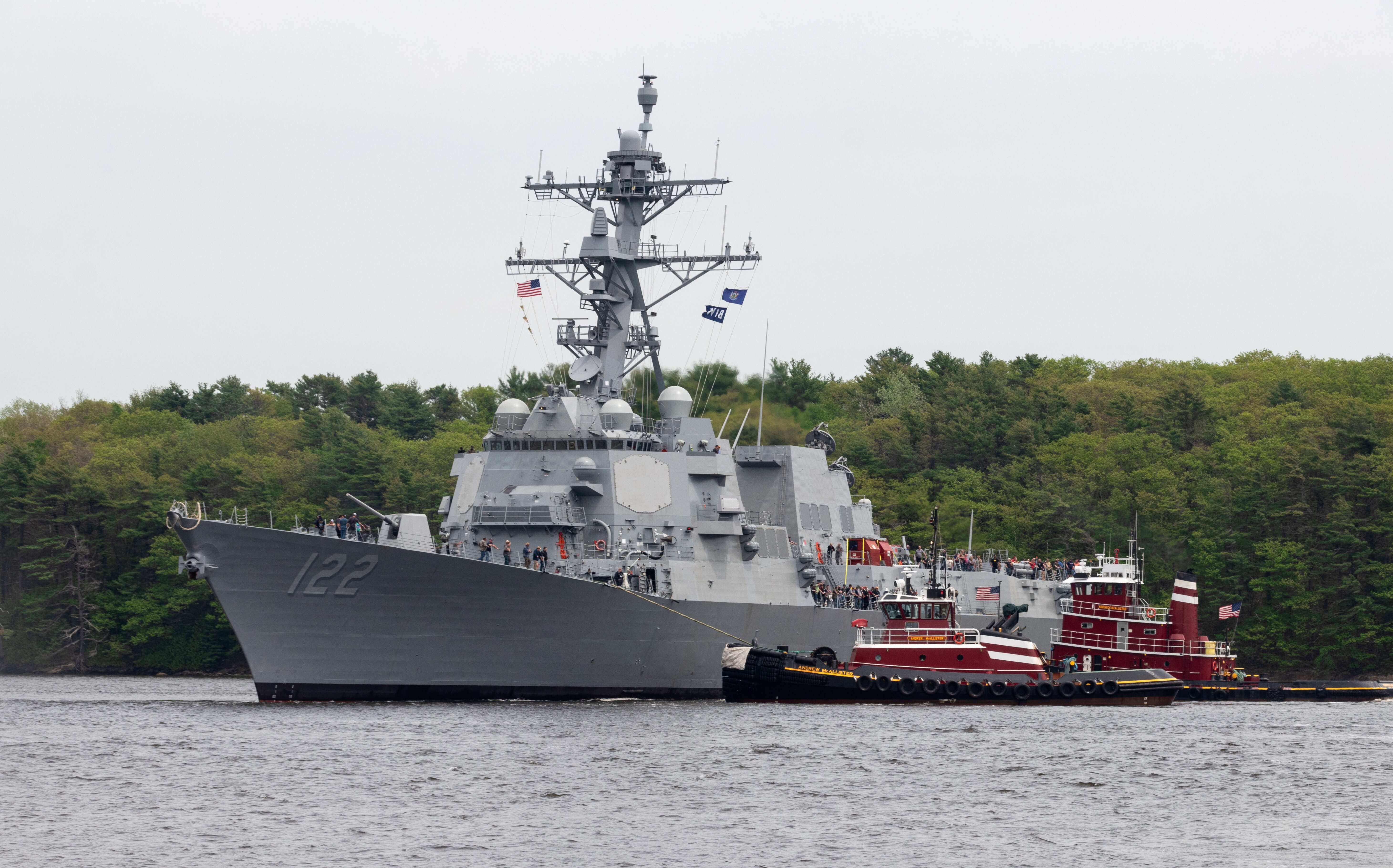 the U.S. Navy's destroyer, built by Bath Iron Works, is flating on the water wtih a couple of tugboats tied up to it.