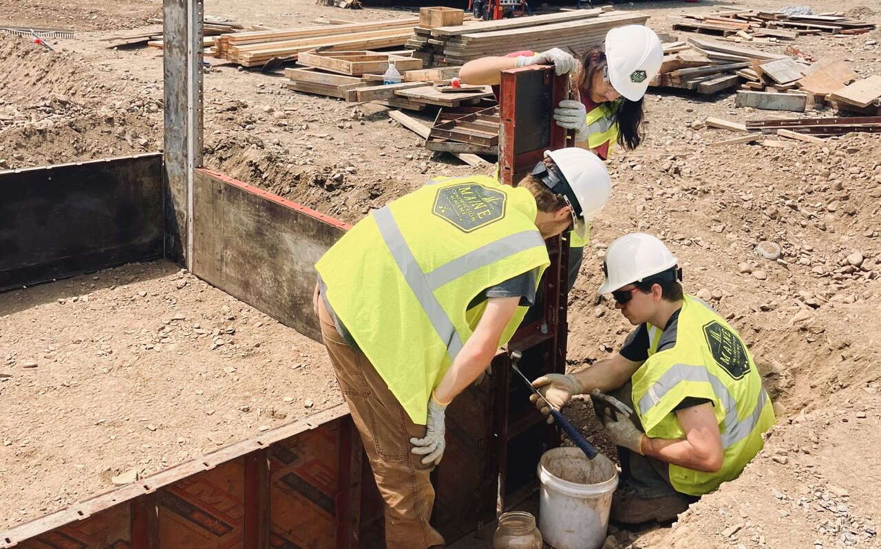 Three students with the Maine Construction Academy learn how to build a foundation. They're looking at tools next to some steel set in the ground.