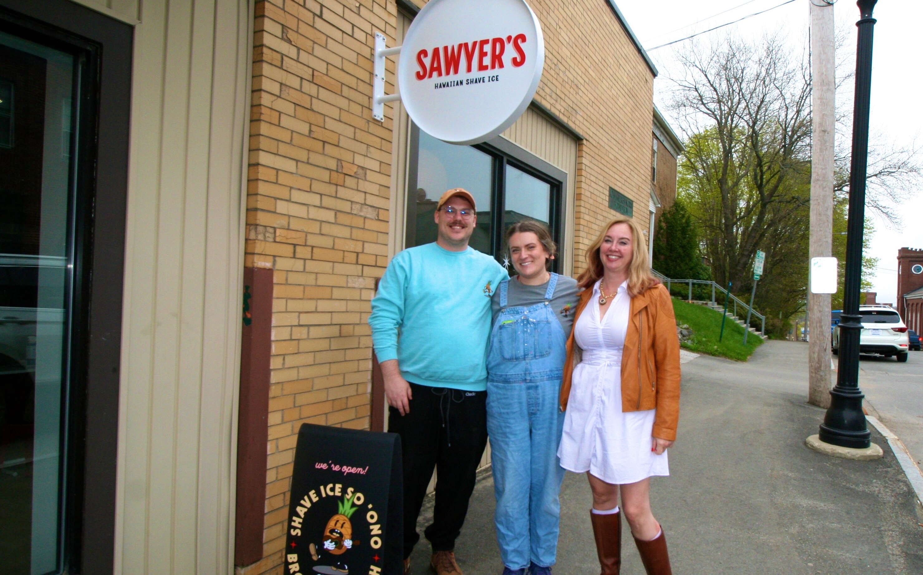 Three people pose on a siewalk with a sign.