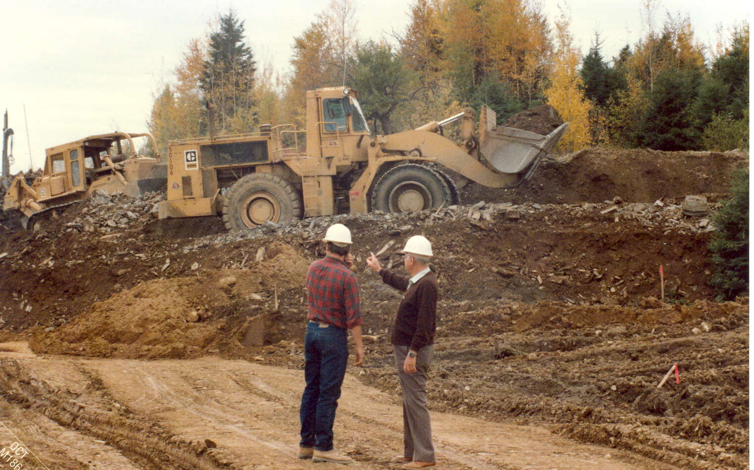 Two people stand outside amidsta lot of bare ground.