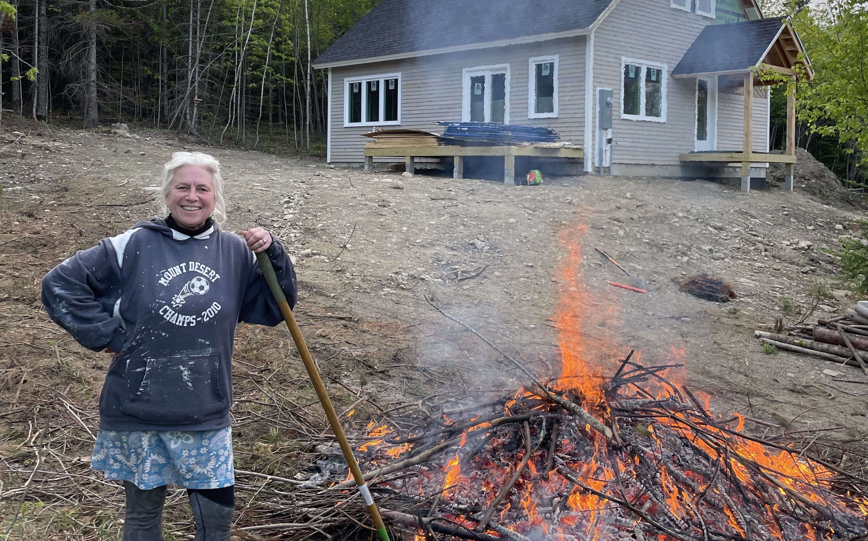 A person stands with a pile of burning brush with a house in the background.