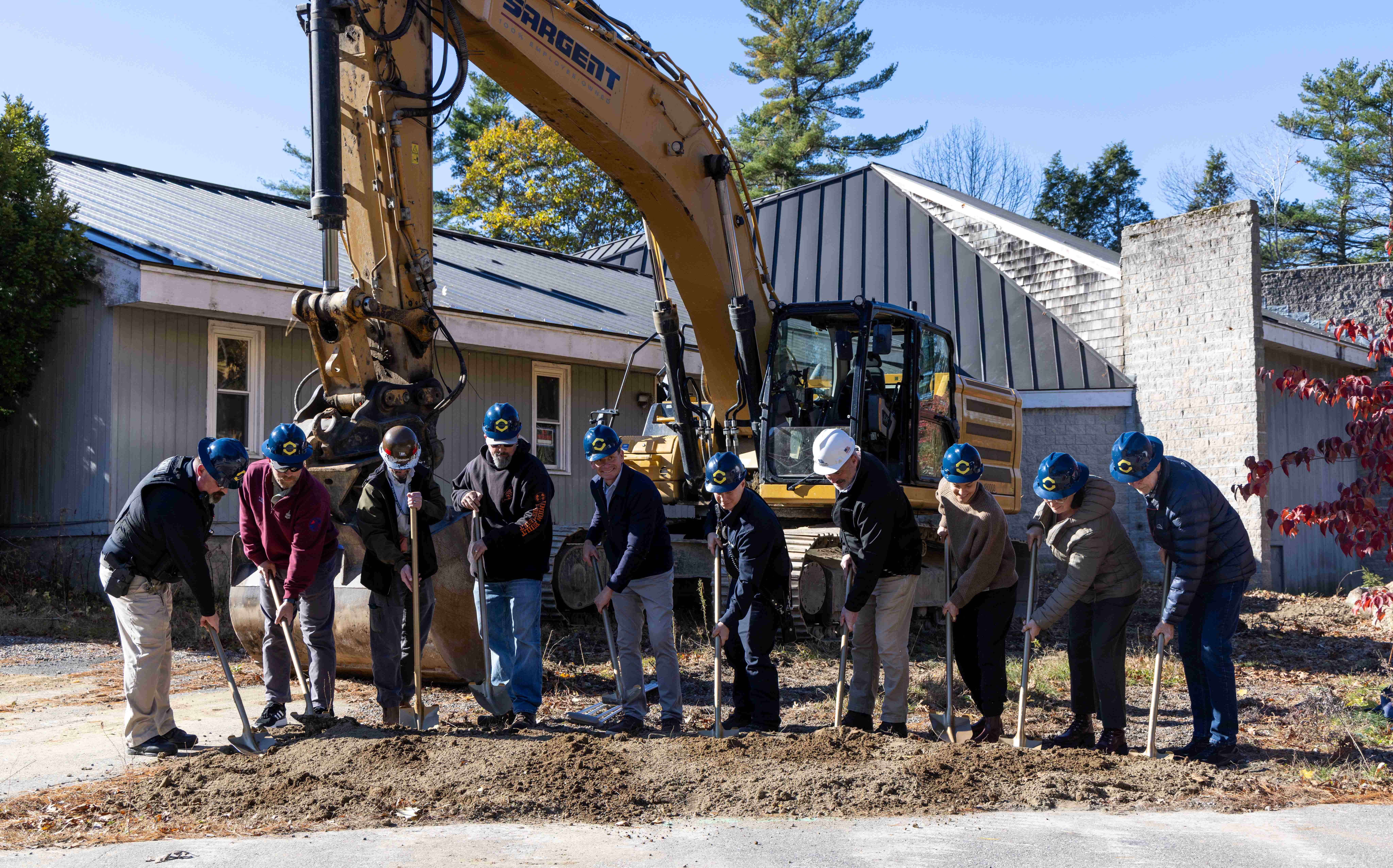 People pose in a line with shovels and an excvatator behind them.